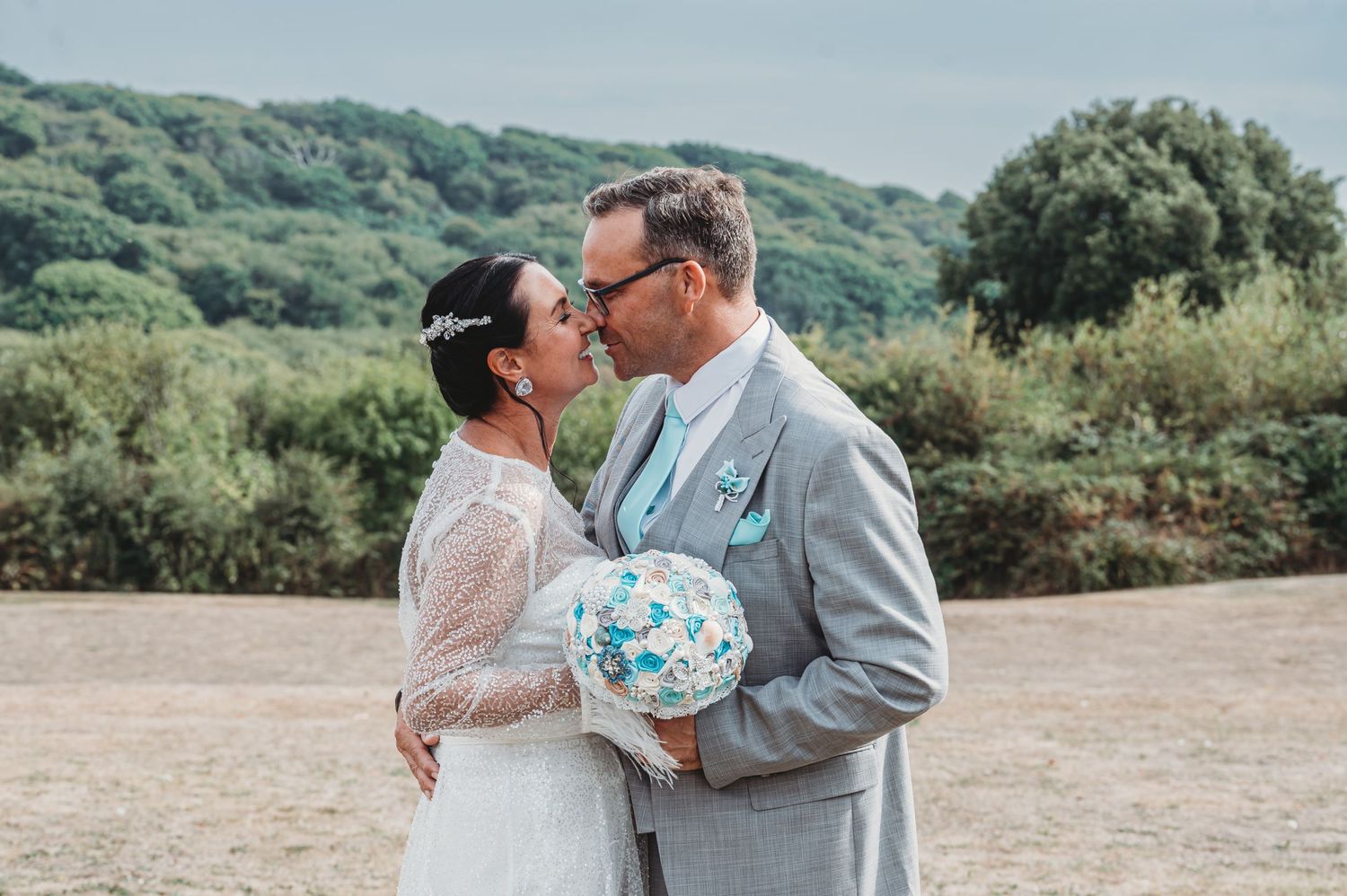 A couple in wedding attire share an intimate moment outdoors, the bride holding a blue and white bouquet.