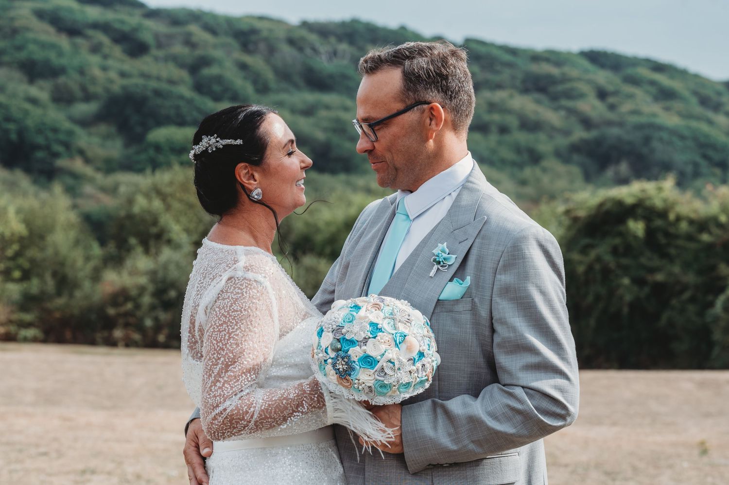 Couple in wedding attire stands close together outdoors holding a blue and white bouquet against a green hillside background.
