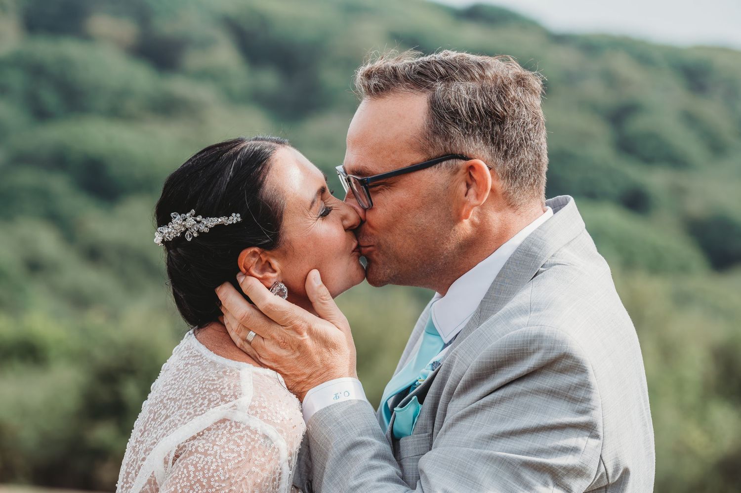 A couple share a romantic kiss on their wedding day against a lush green mountainous backdrop.