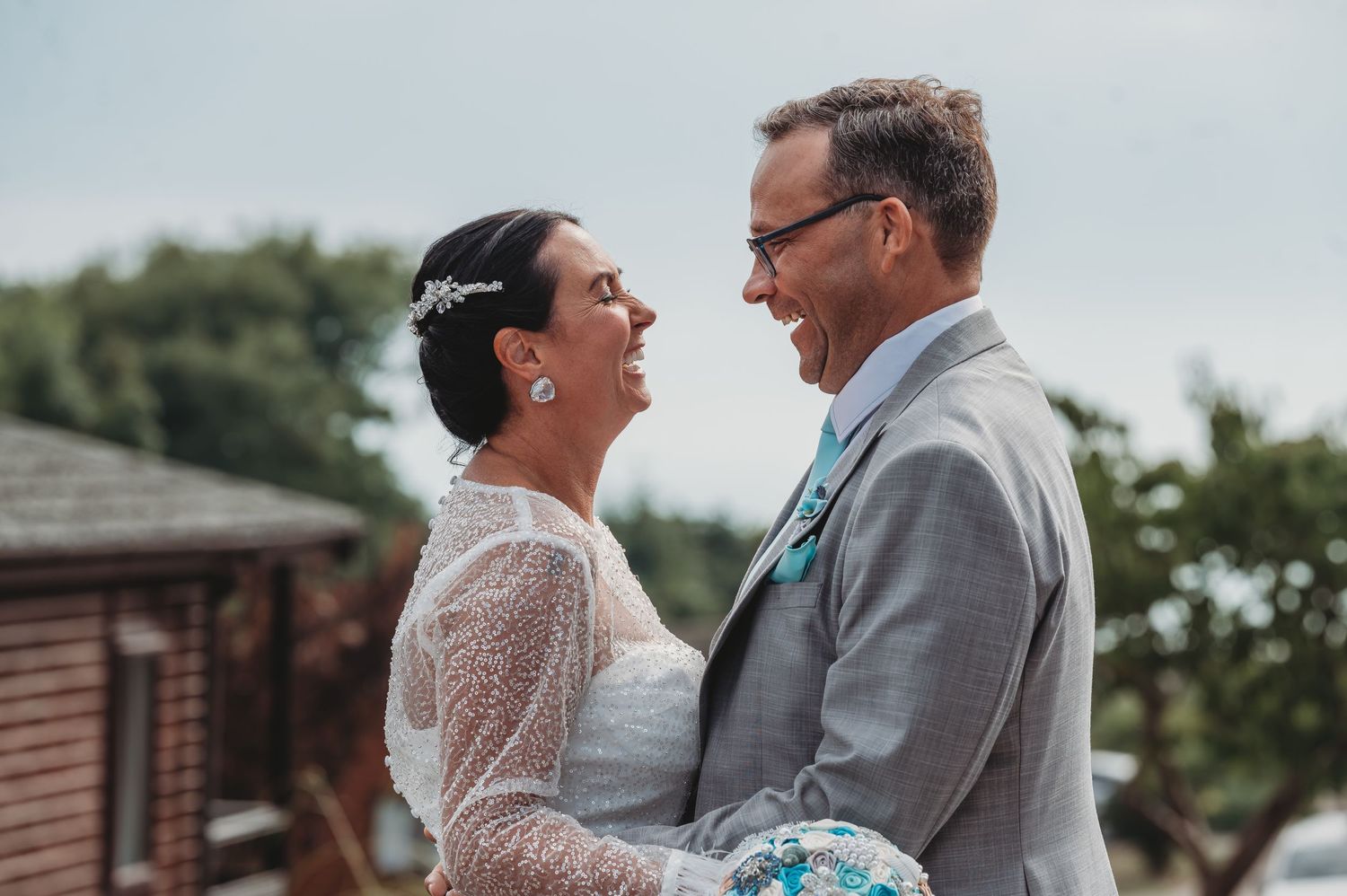 A couple shares a joyful moment together outdoors on their wedding day, the bride in lace and groom in gray suit.