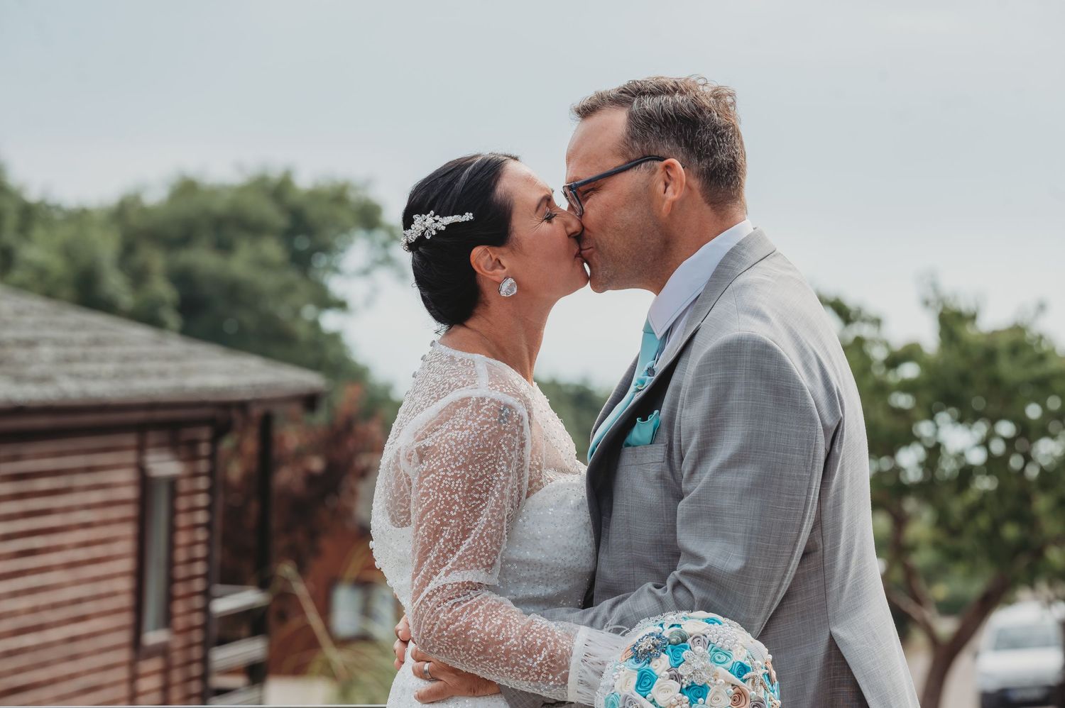 A romantic wedding moment outdoors with the couple sharing a kiss while the bride holds a blue and white bouquet.