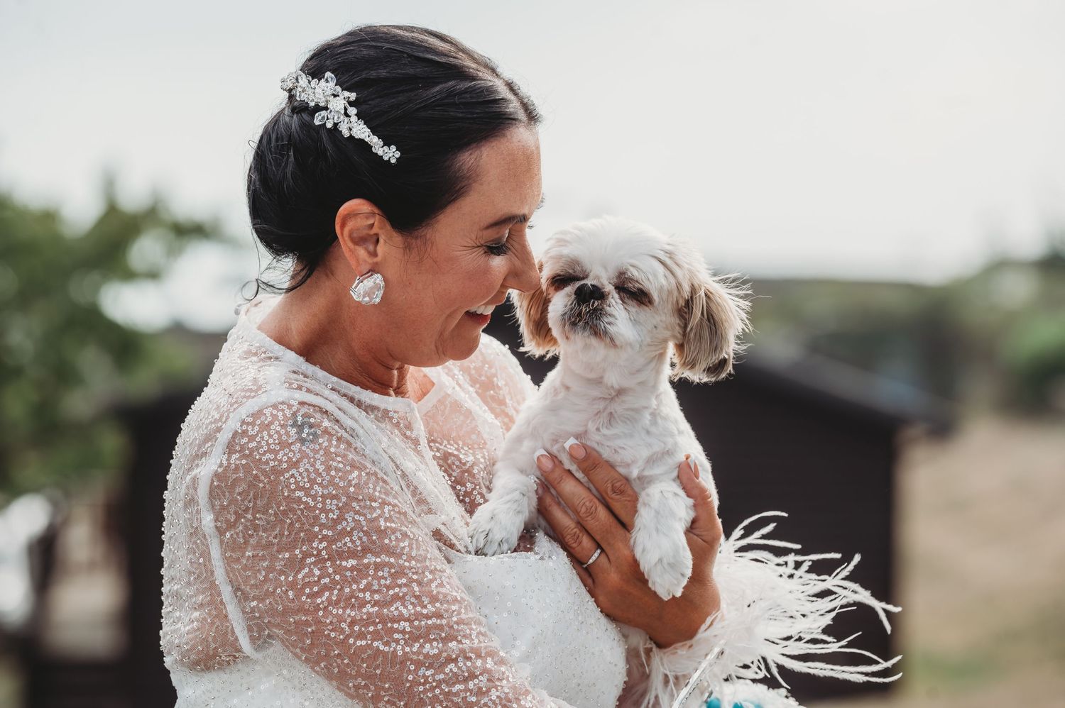 A bride in a lace wedding dress shares a joyful moment with her small white dog on her wedding day.
