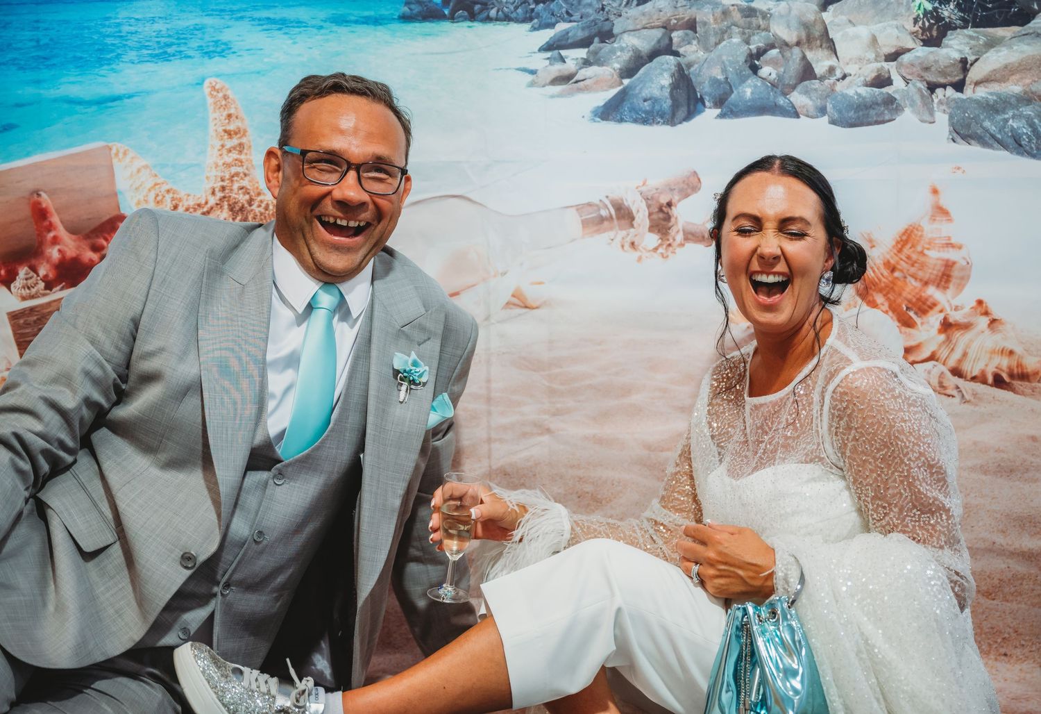 A joyful wedding couple shares a laugh while sitting on a beach with turquoise water and rocky outcrops in the background.