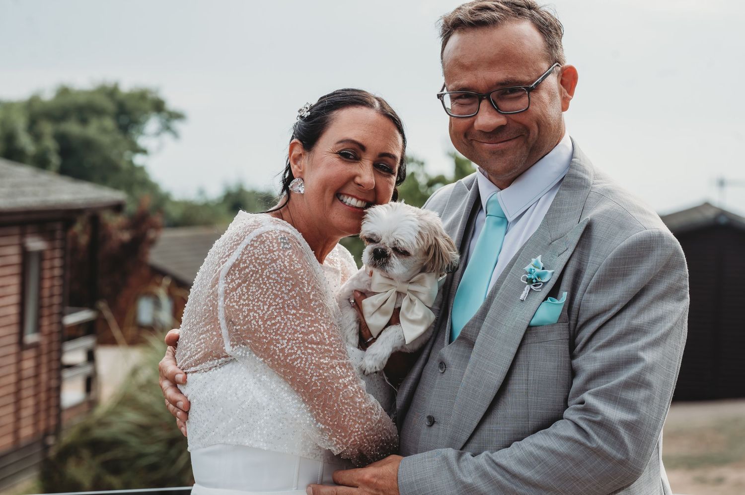 A couple in wedding attire holds their small white dog while posing together outdoors.