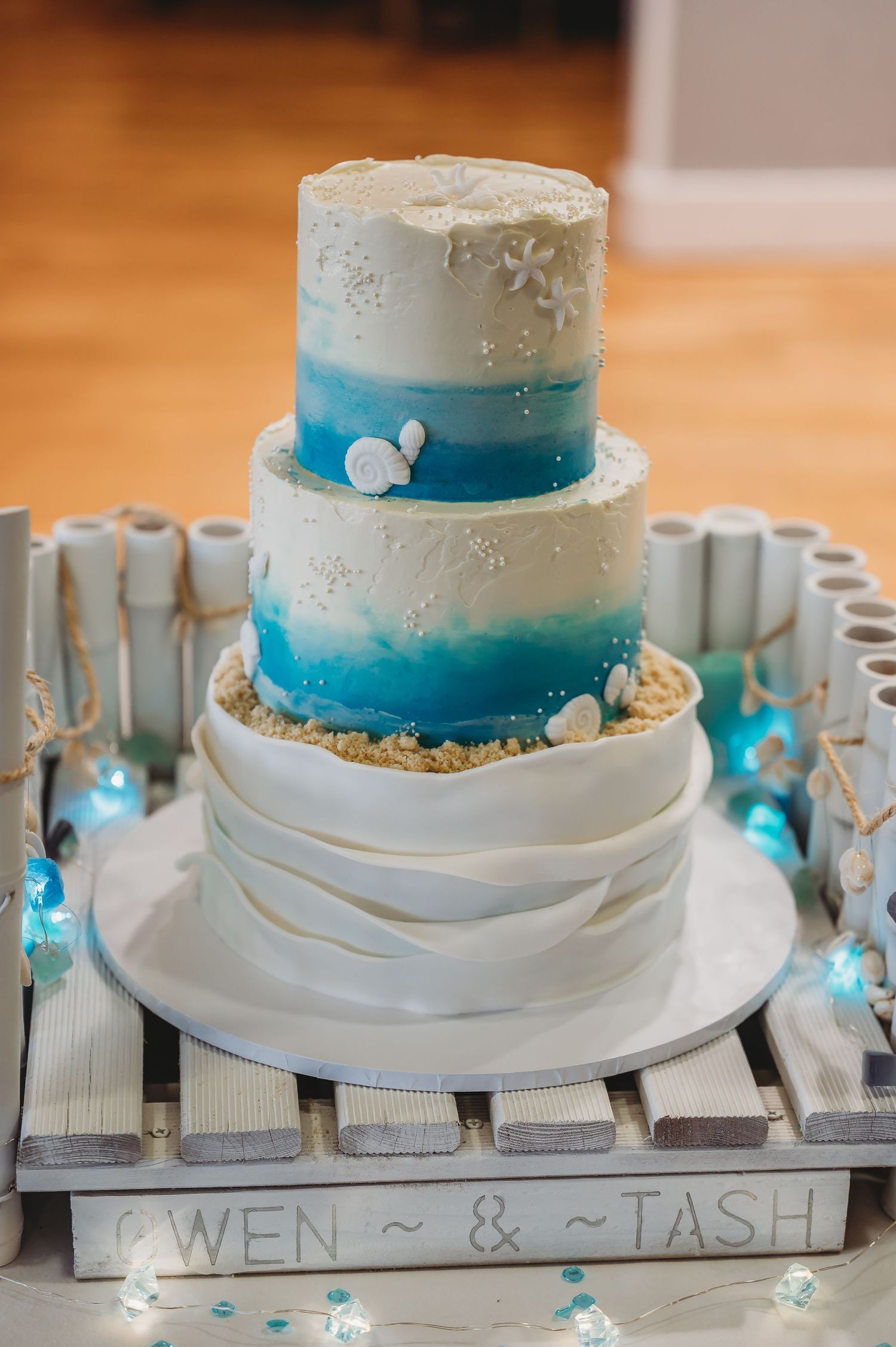 Three-tier beach themed wedding cake with white and turquoise ombre design on a wooden pallet display with twinkle lights.