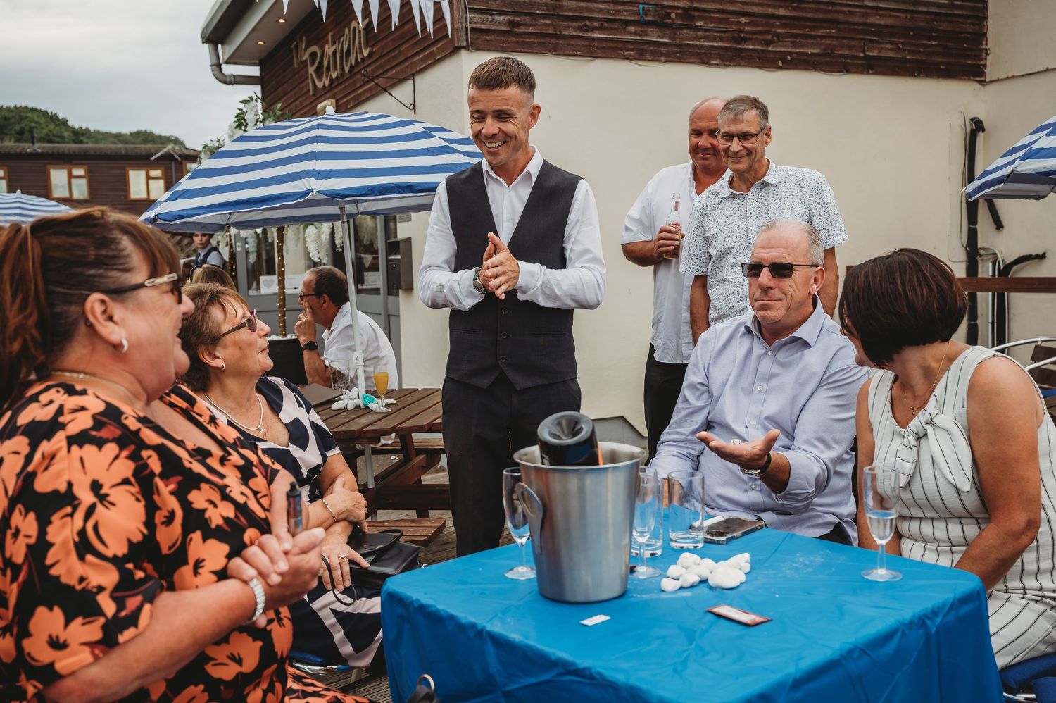 A group of people gathered around a blue tablecloth-covered table with a beverage dispenser outdoors at a social event.