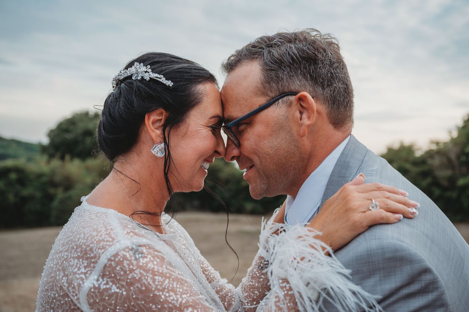 A couple share an intimate moment during their wedding, dressed in formal attire against an outdoor backdrop.