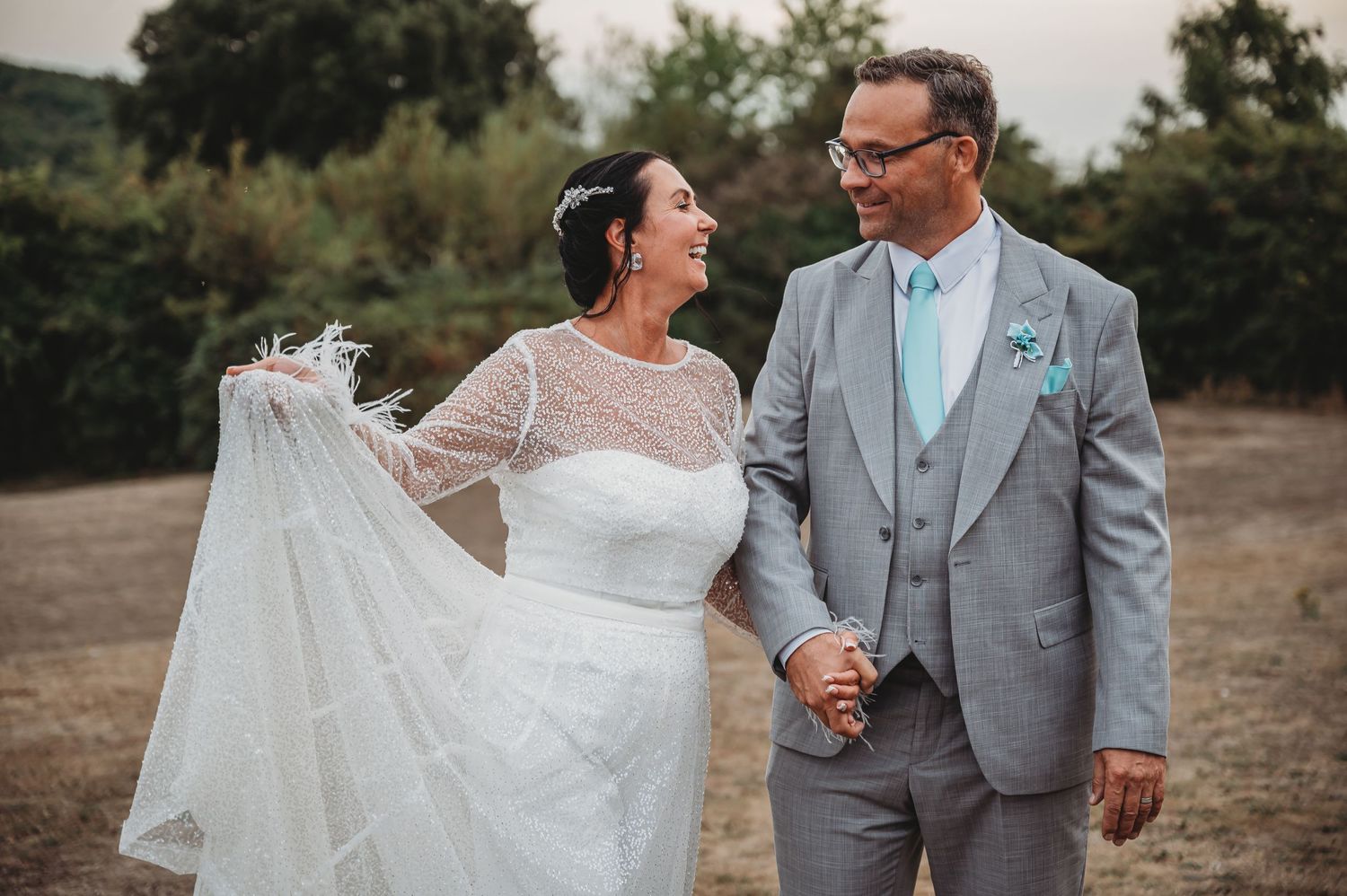 A radiant bride in white lace dress and groom in grey suit share a joyful moment outdoors.