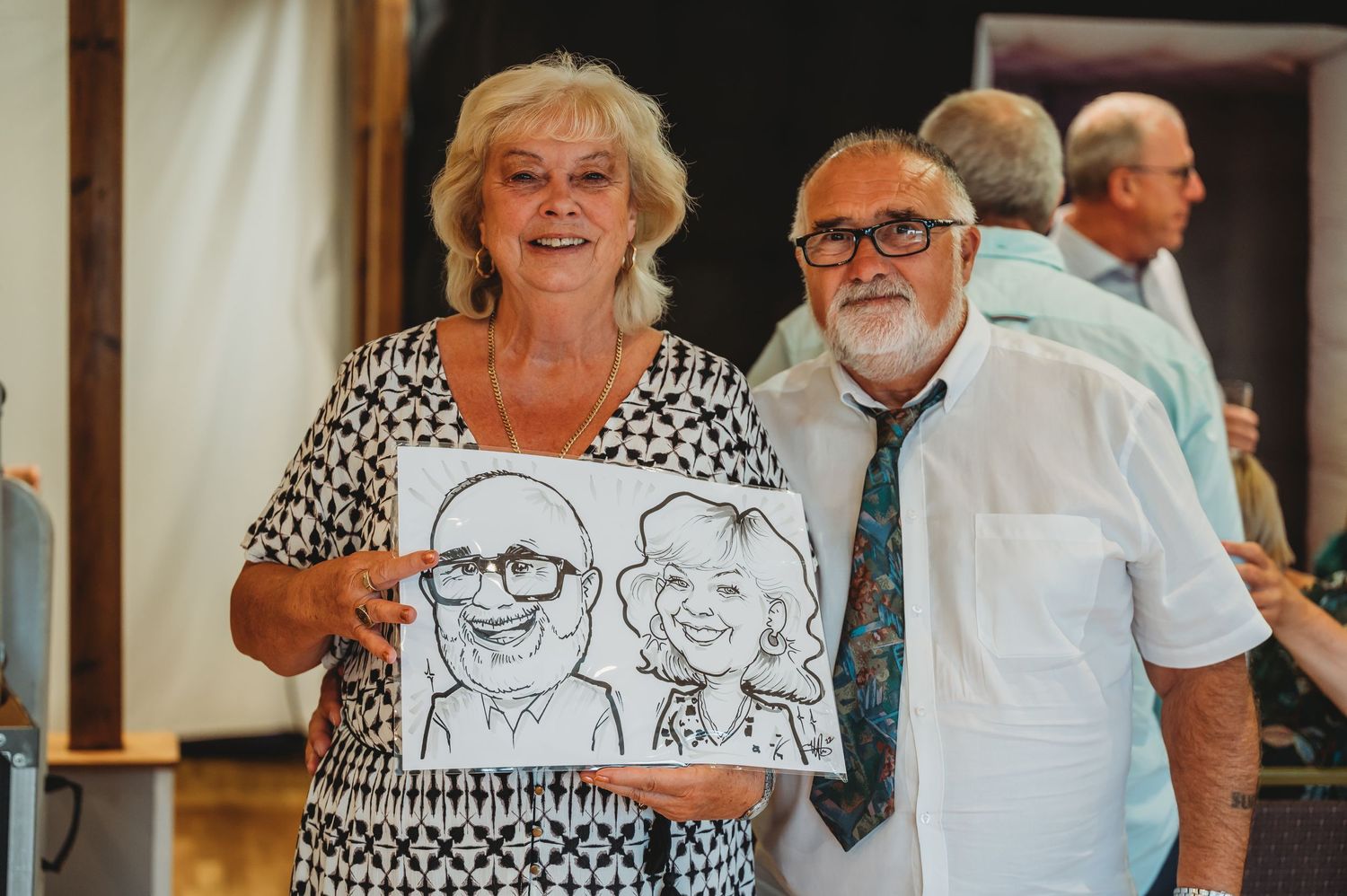 A couple poses with their black and white portrait sketch, with the woman wearing a polka dot top and smiling for the camera.