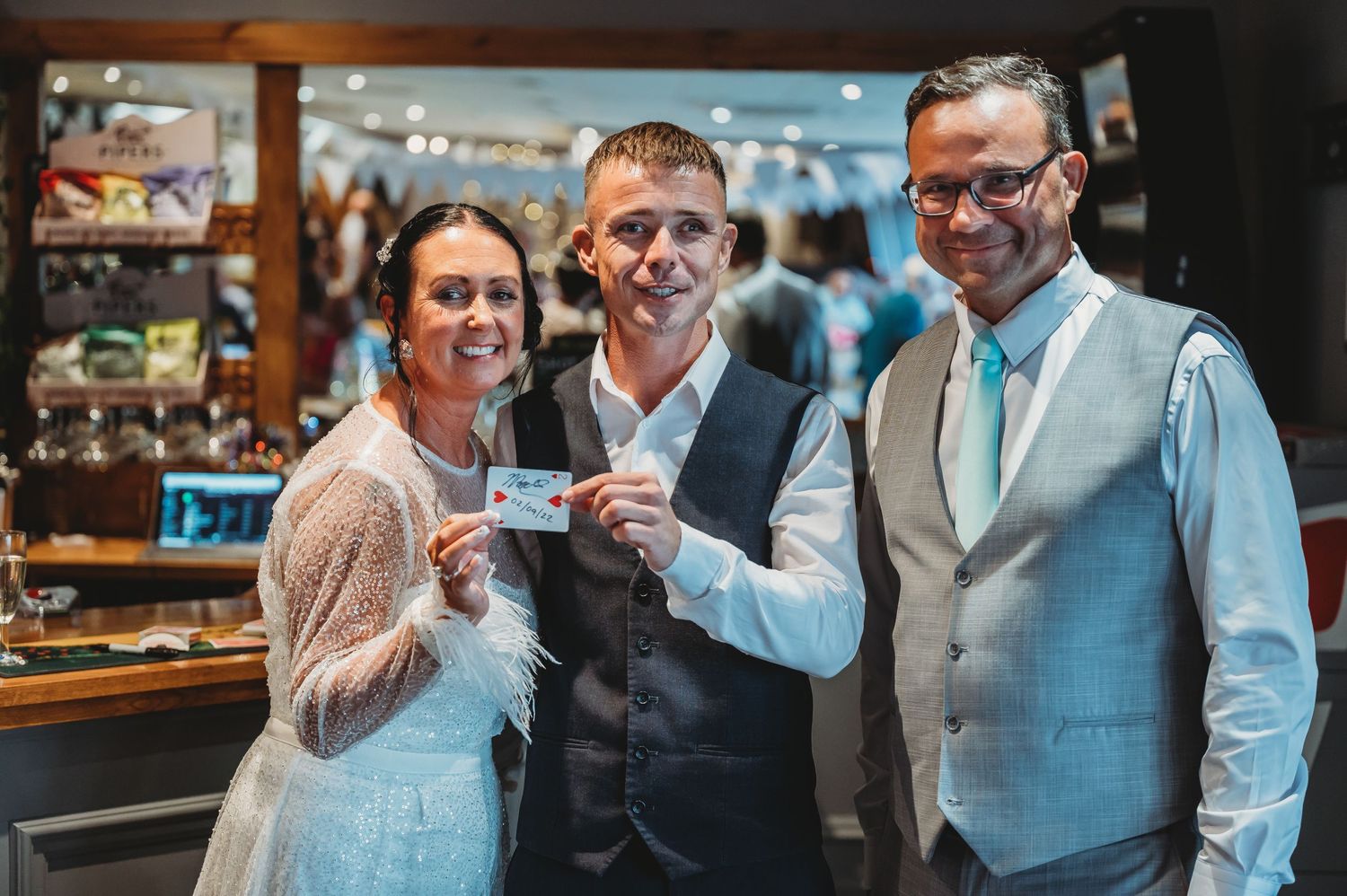 Three people stand smiling together at a bar counter holding small cards or tickets in their hands.