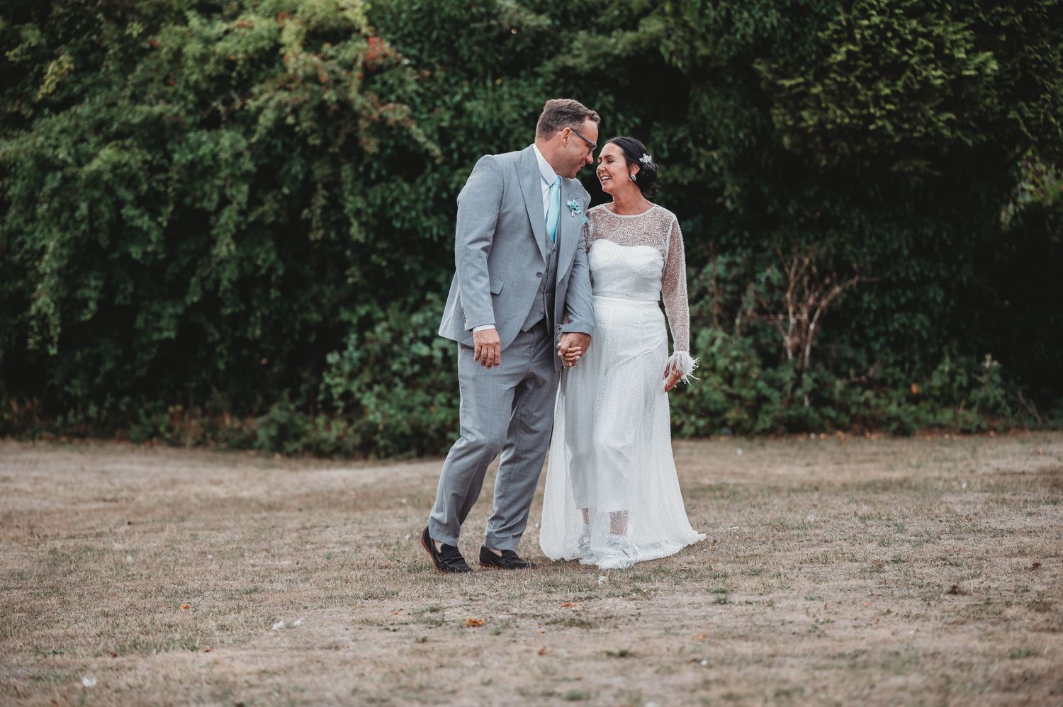 A couple in wedding attire share a tender moment together while standing on a dirt path near green foliage.