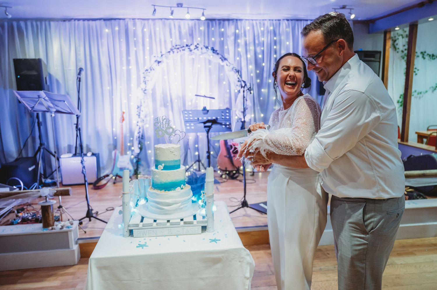 A couple shares a joyful moment while cutting their blue and white tiered wedding cake at an indoor reception.