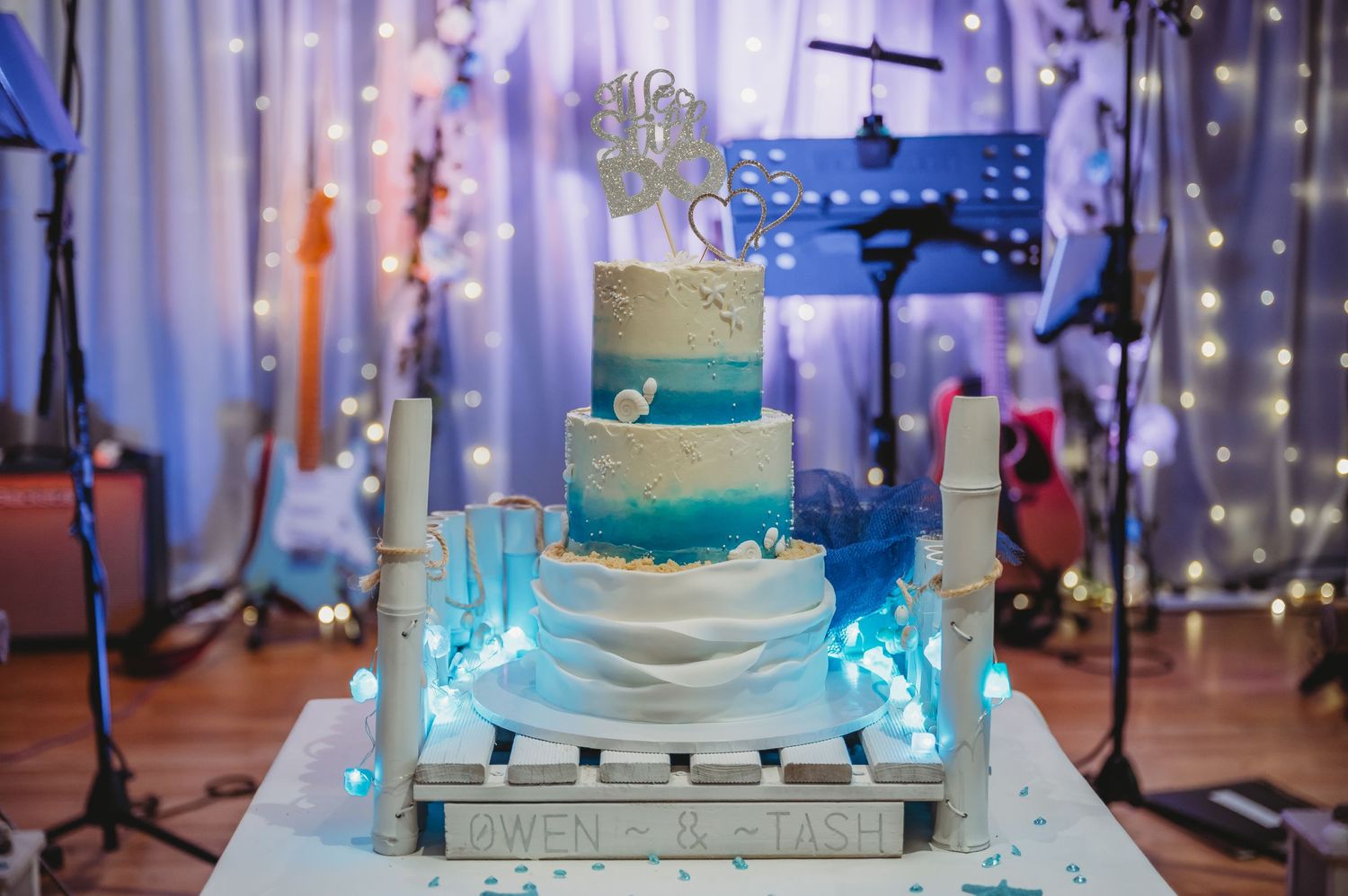 An elegant four-tier ombre wedding cake in shades of turquoise and white sits on a lit platform with sparkly backdrop.