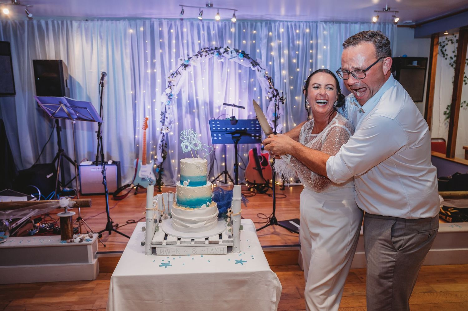 A couple cuts their teal and white wedding cake together under a decorative arch with string lights at an indoor reception.