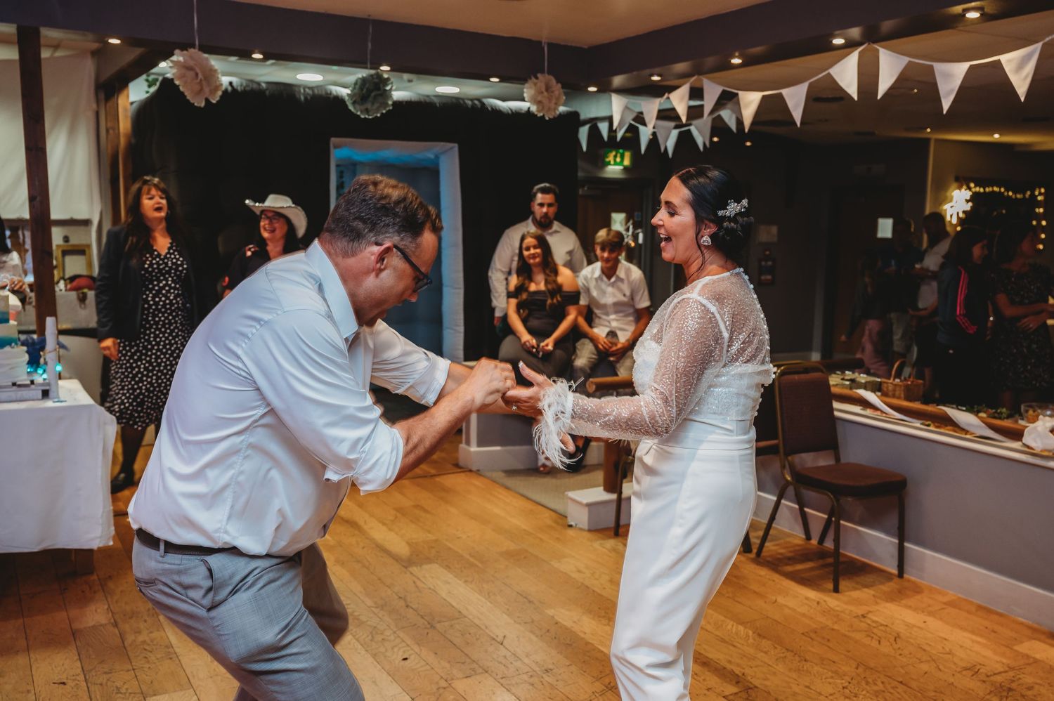 A couple dances joyfully together on a wooden floor at a wedding reception decorated with bunting and lights.