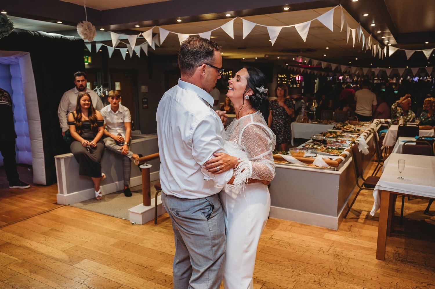 A couple shares their first dance on a wooden floor at a wedding reception decorated with white bunting and string lights.