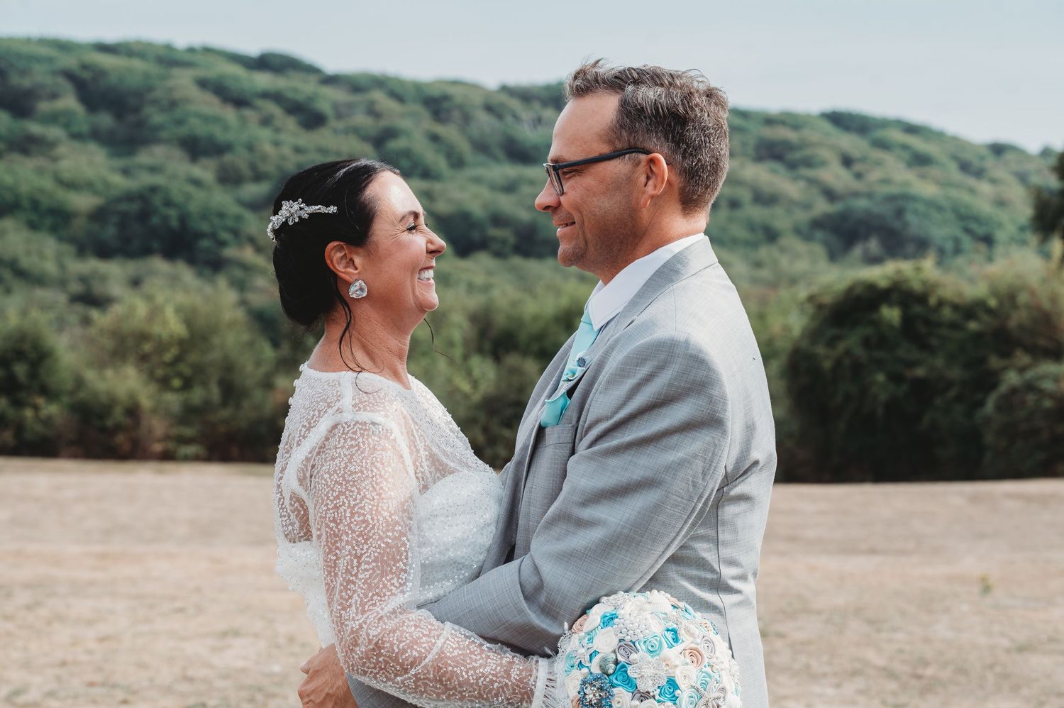A couple in wedding attire share a joyful moment together outdoors against a green hillside landscape.