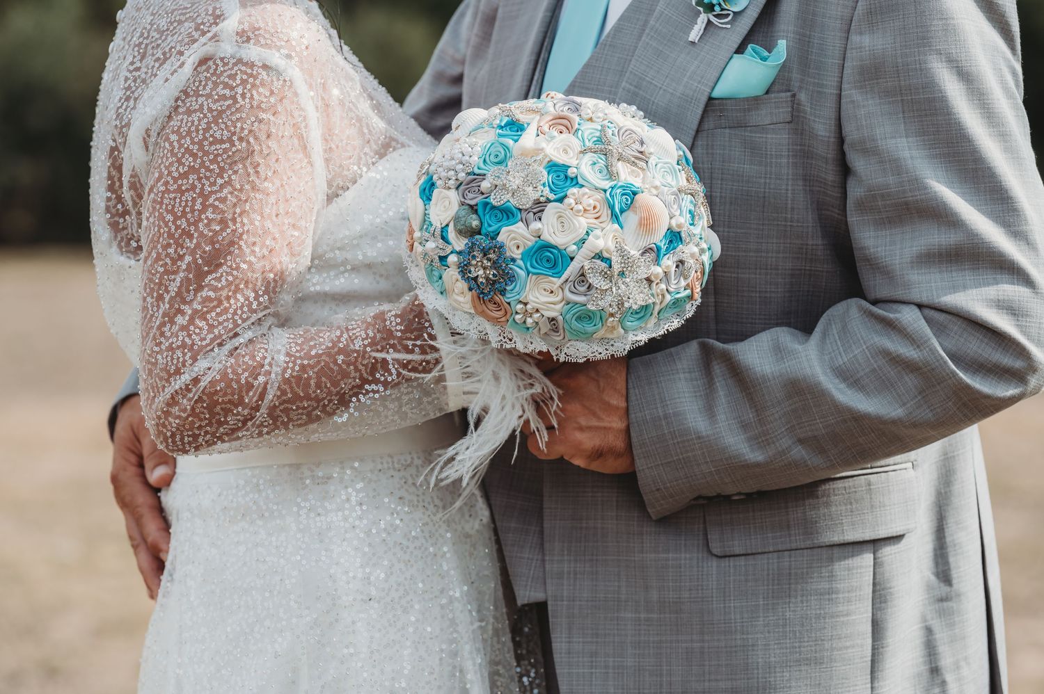 Close-up of a wedding couple holding a unique turquoise and white brooch bouquet against a lace dress and grey suit.