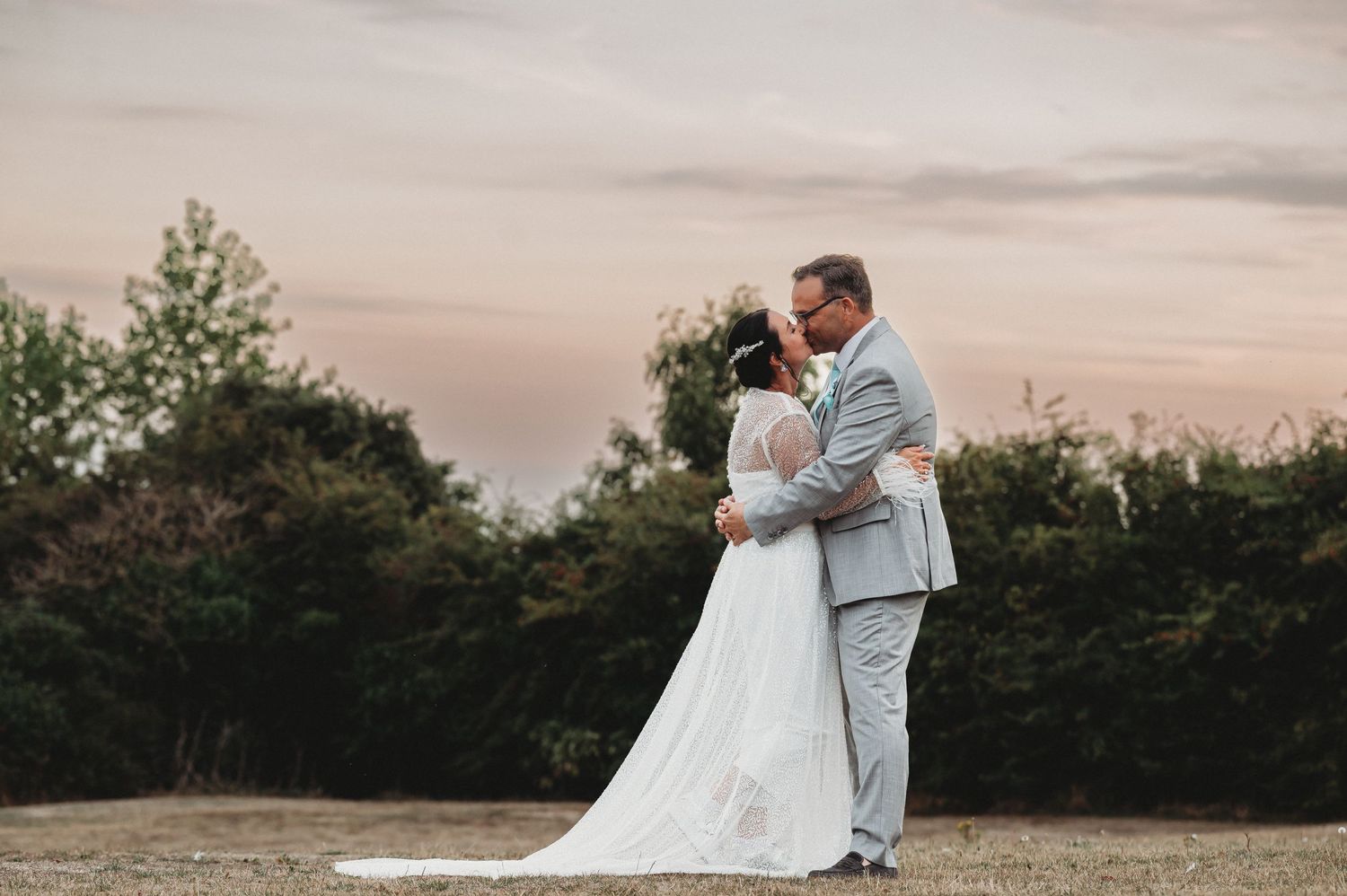 A couple in wedding attire embraces on a hilltop at sunset with trees in the background.