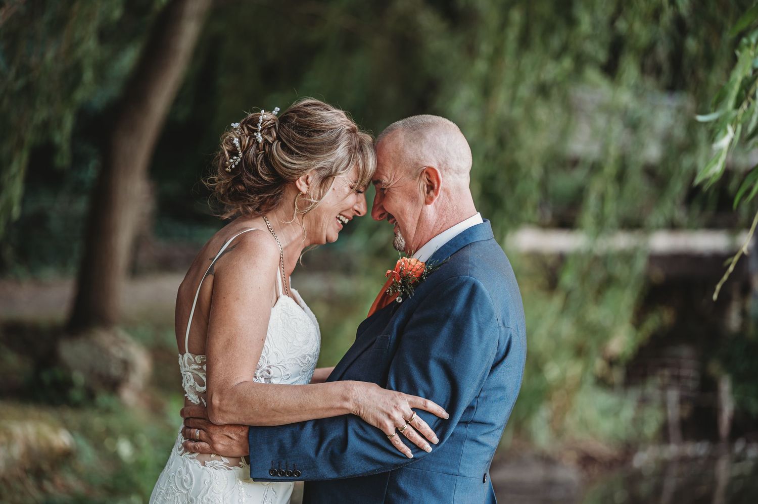 A romantic wedding couple shares an intimate moment outdoors with trees and soft lighting in the background.