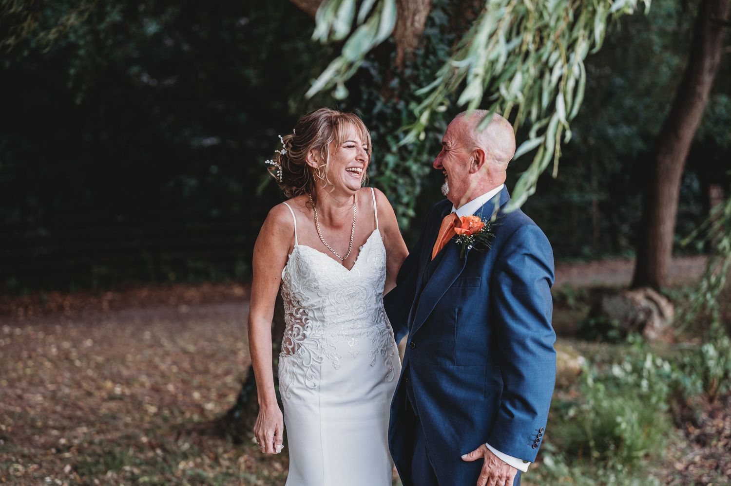 A couple shares a candid moment under willow trees during their wedding celebration, wearing formal navy suit and white dress.