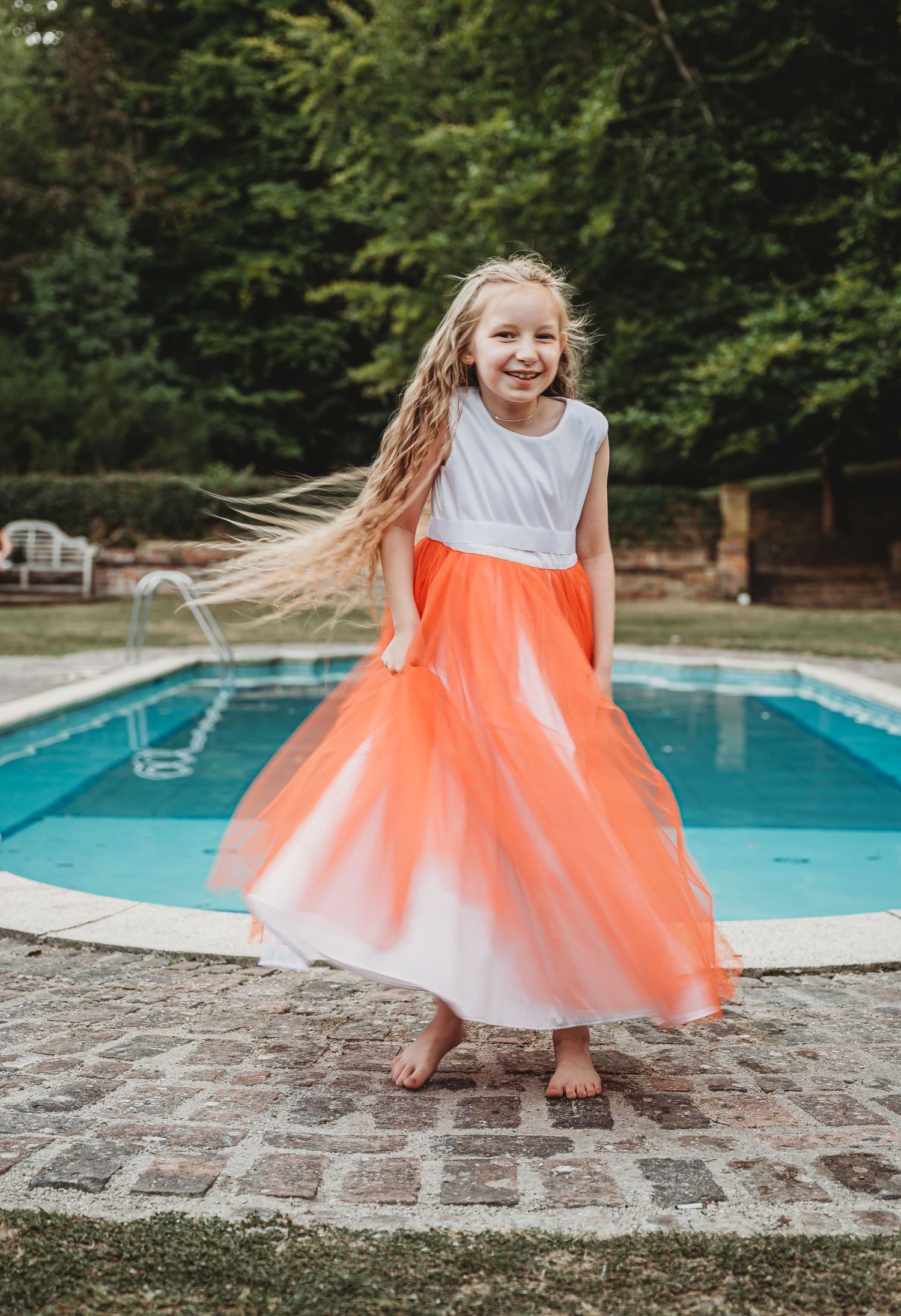 Young dancer in white and coral tulle dress twirls joyfully by a swimming pool surrounded by trees.