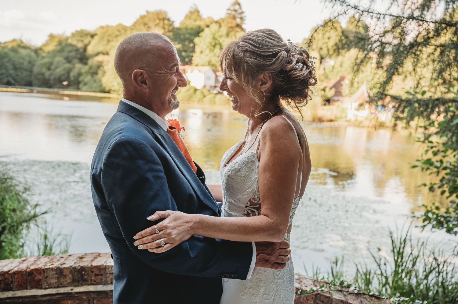 A couple in wedding attire embraces by a scenic lake during golden hour, creating a romantic waterfront moment.