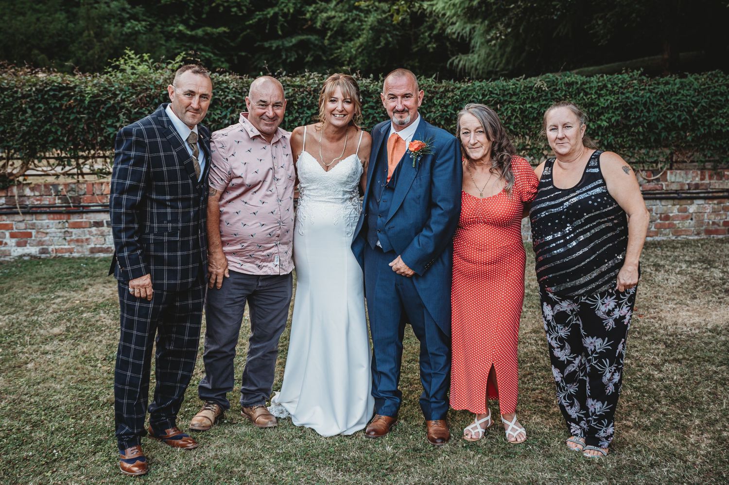 A group of wedding guests pose together outdoors against a brick wall on a sunny day.