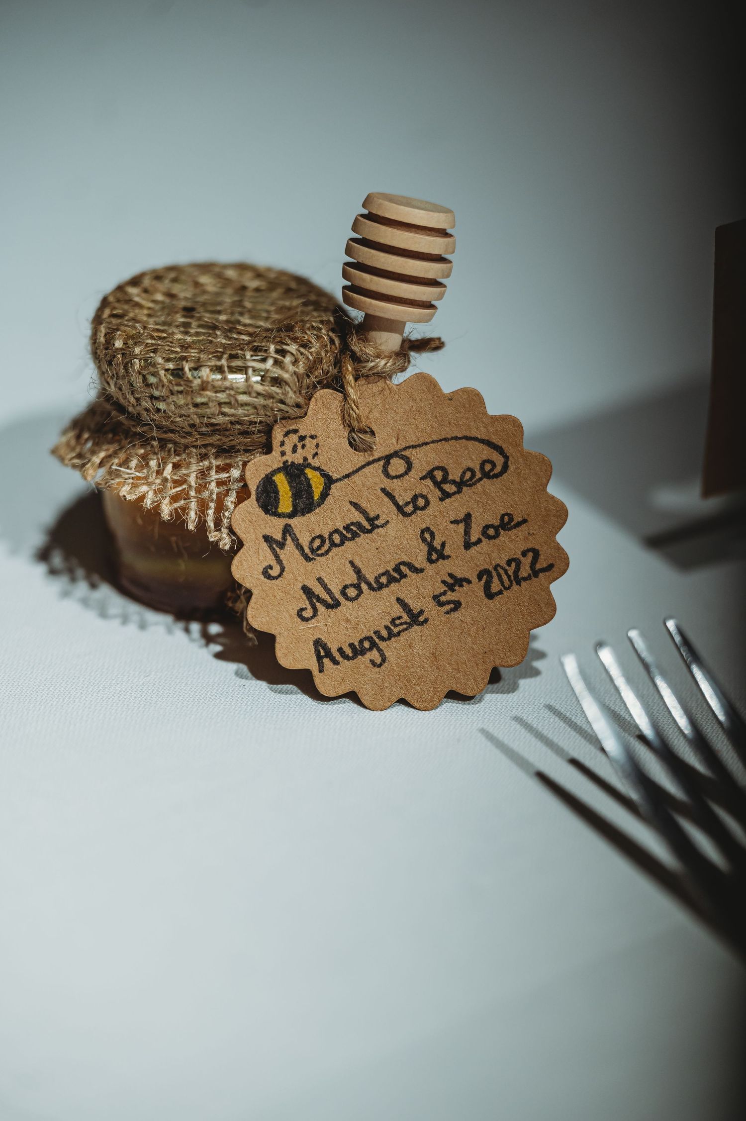 A small jar of honey with a rustic burlap top and decorative bee tag labeled "Meant to bee" as a wedding favor from August 2022.
