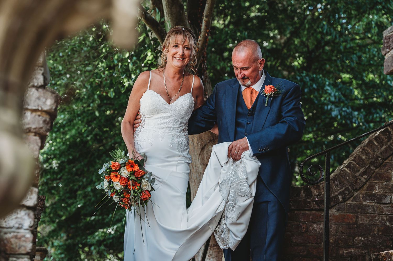 A couple on their wedding day walk together in a garden setting with the bride holding a vibrant orange bouquet.