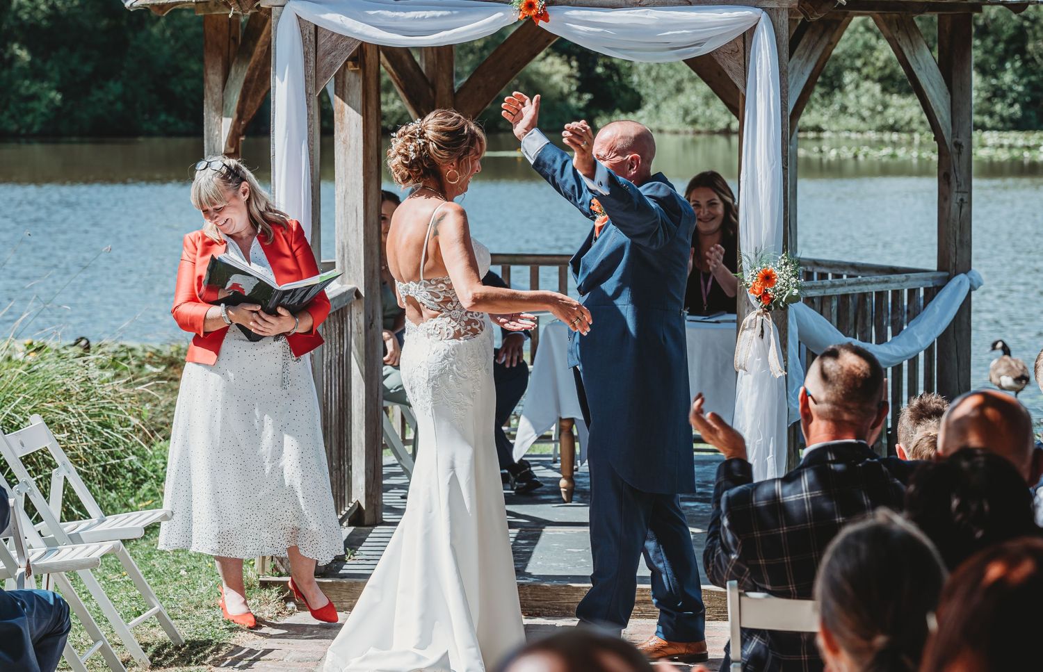 A lakeside wedding ceremony takes place under a decorated gazebo with white fabric draping and guests seated outdoors.