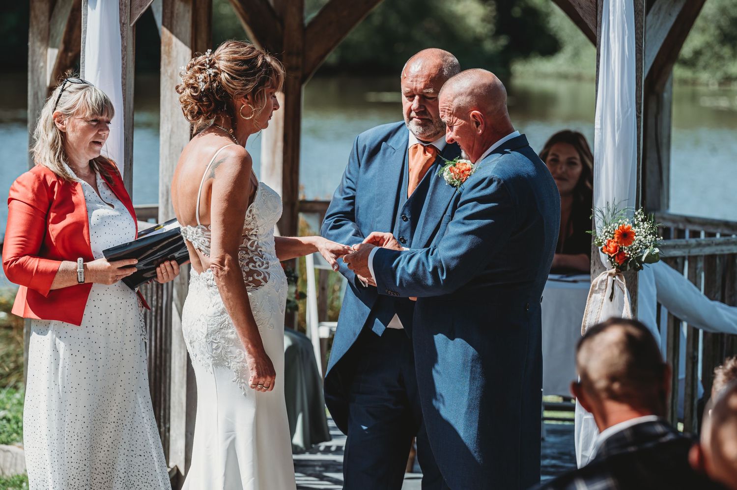 An outdoor lakeside wedding ceremony takes place on a deck with people gathered in formal attire.