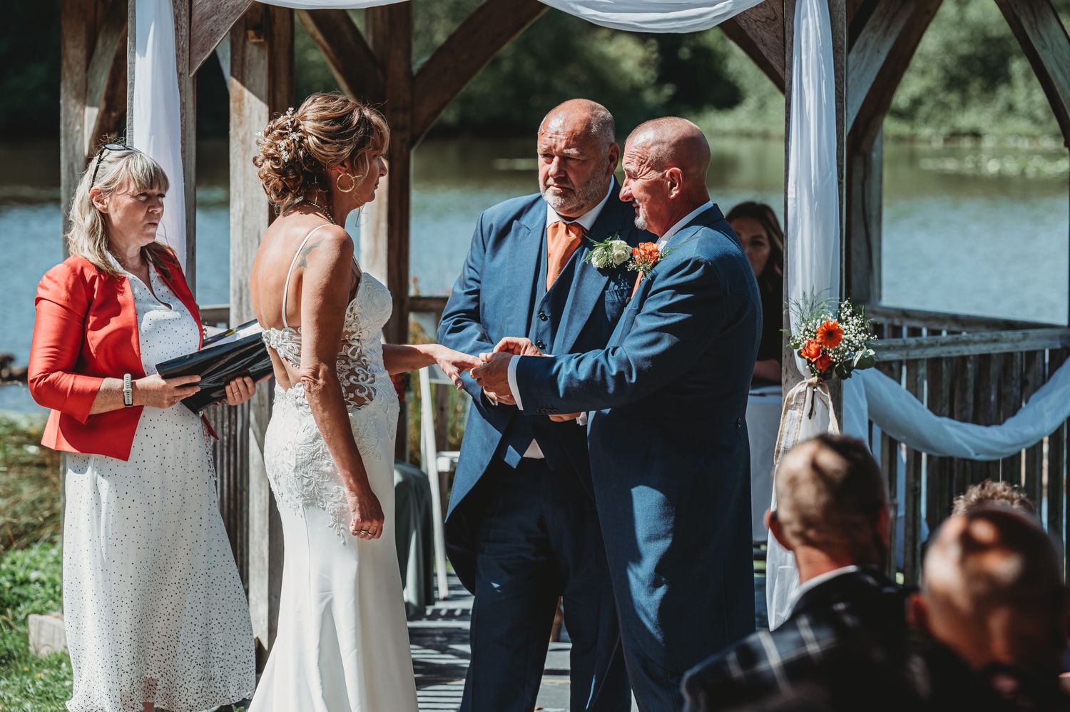 Outdoor lakeside wedding ceremony takes place under a covered pavilion on a sunny summer day.
