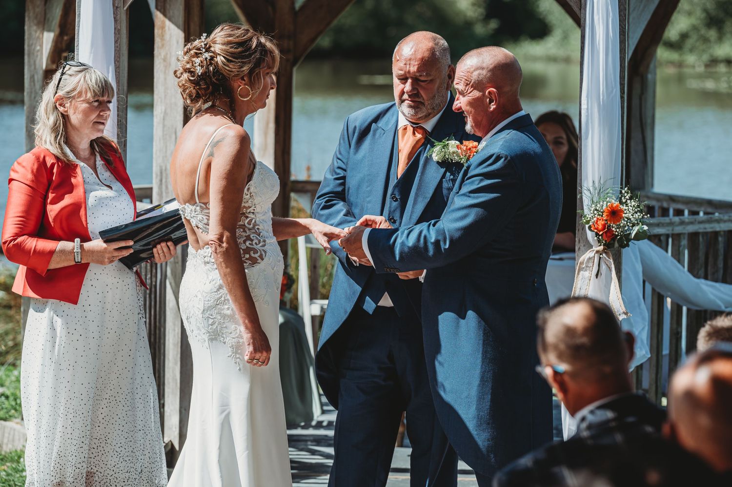 Wedding ceremony taking place on a covered porch overlooking a lake with an officiant reading from a small book.