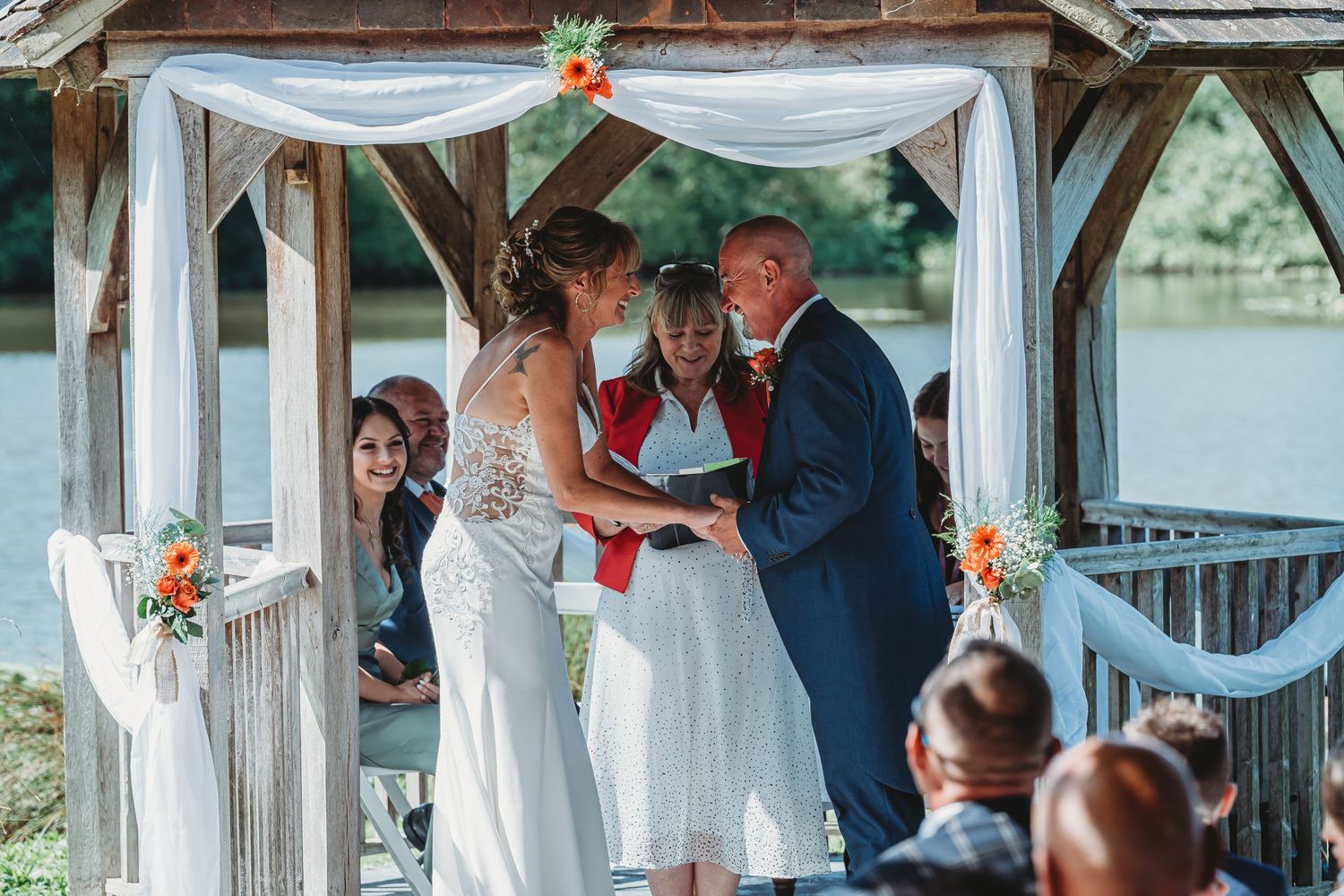 An outdoor wedding ceremony takes place under a decorated white gazebo by a serene lake.