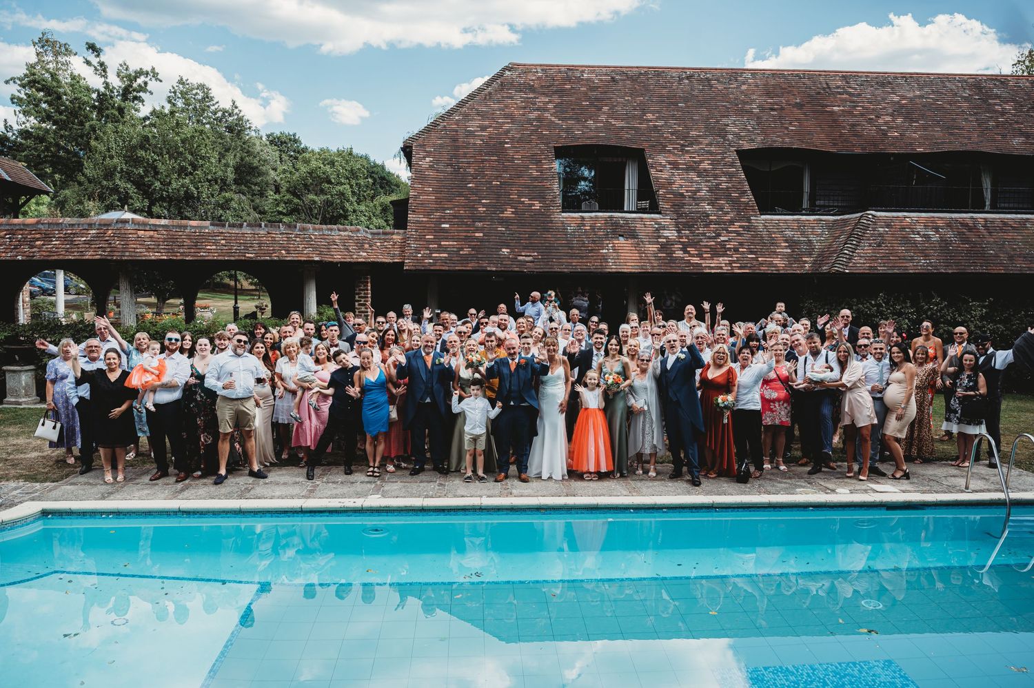 A large group of wedding attendees gather for a photo beside a swimming pool at a rustic venue on a sunny day.