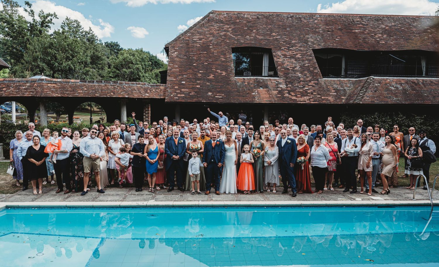 Large group of wedding guests gathered for a group photo beside an outdoor swimming pool at a rustic venue.