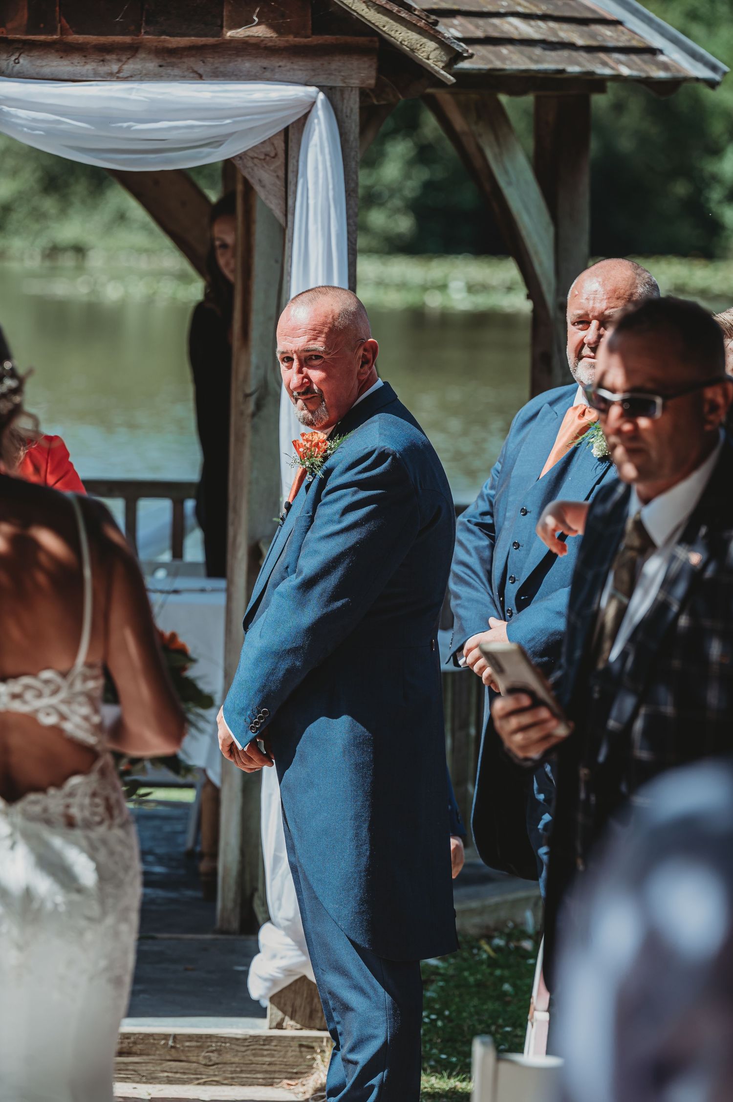 Wedding party members in blue suits stand under a decorated gazebo by a lake during an outdoor ceremony.