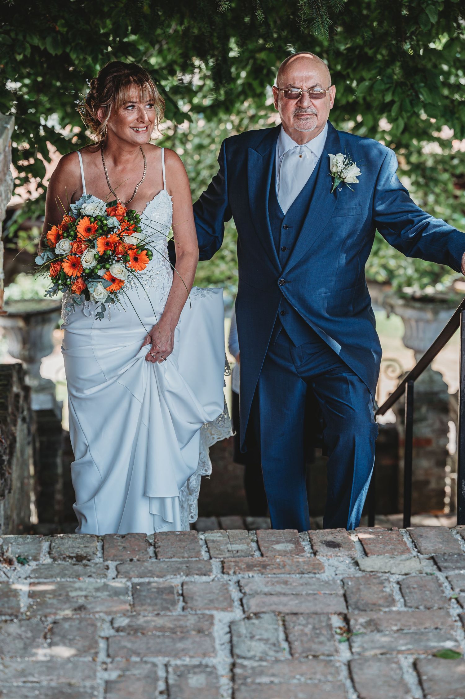 A newly married couple walks together on brick steps, the bride in white holding orange flowers and groom in navy suit.