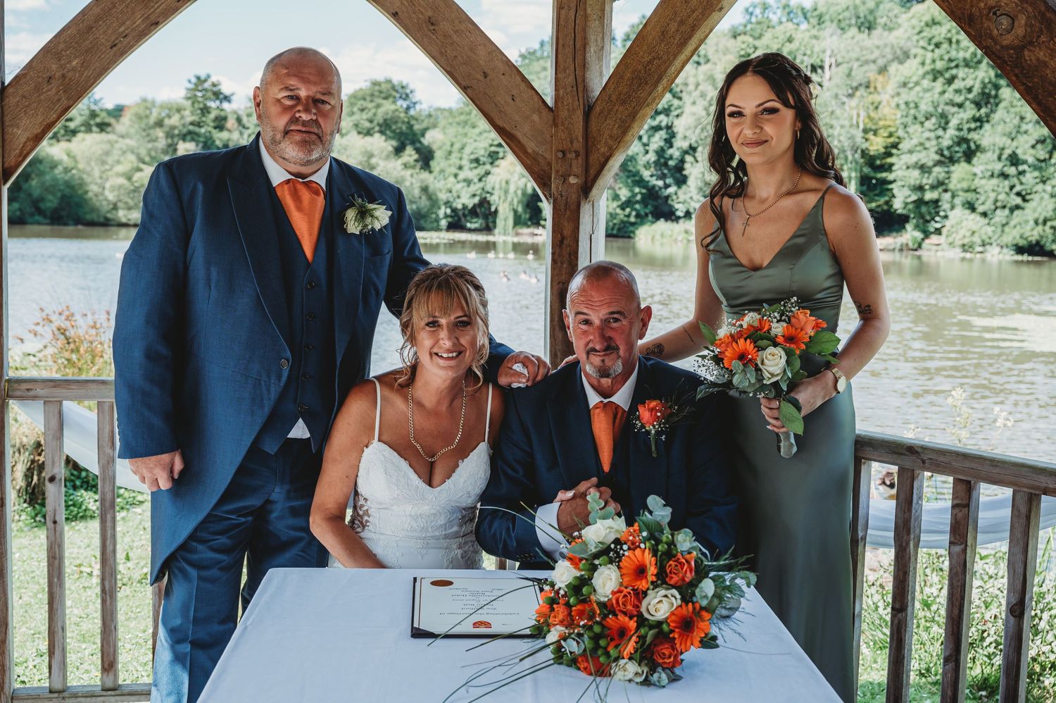 A wedding party gathers at a lakeside gazebo with orange floral bouquets and formal attire during a signing ceremony.