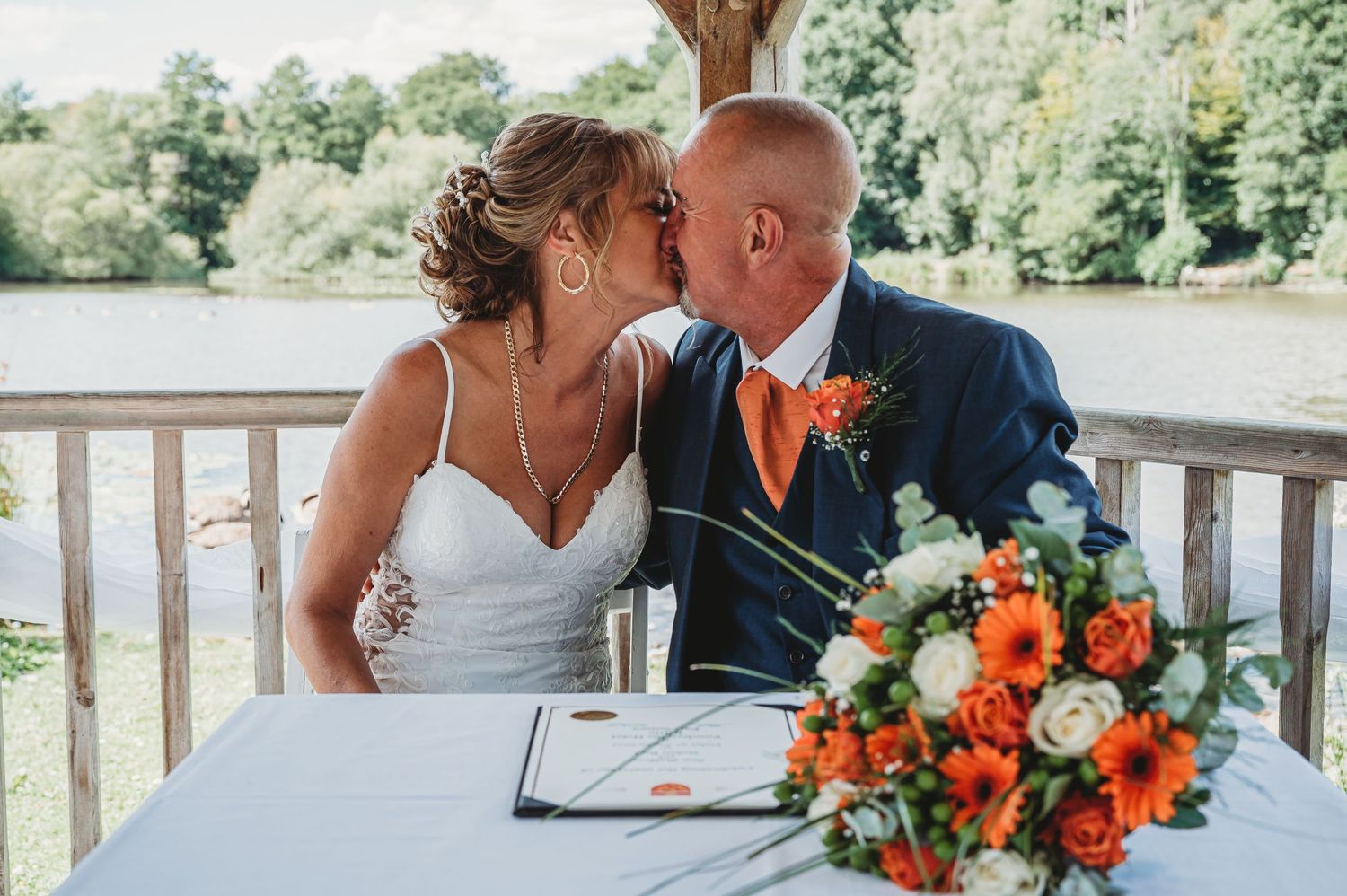 A newlywed couple shares a kiss at a table with vibrant orange and white floral arrangements on their wedding day.