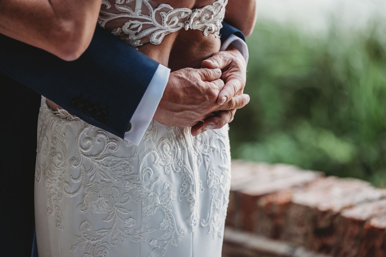 Close-up view of hands holding onto elegant lace wedding dress against green blurred background.
