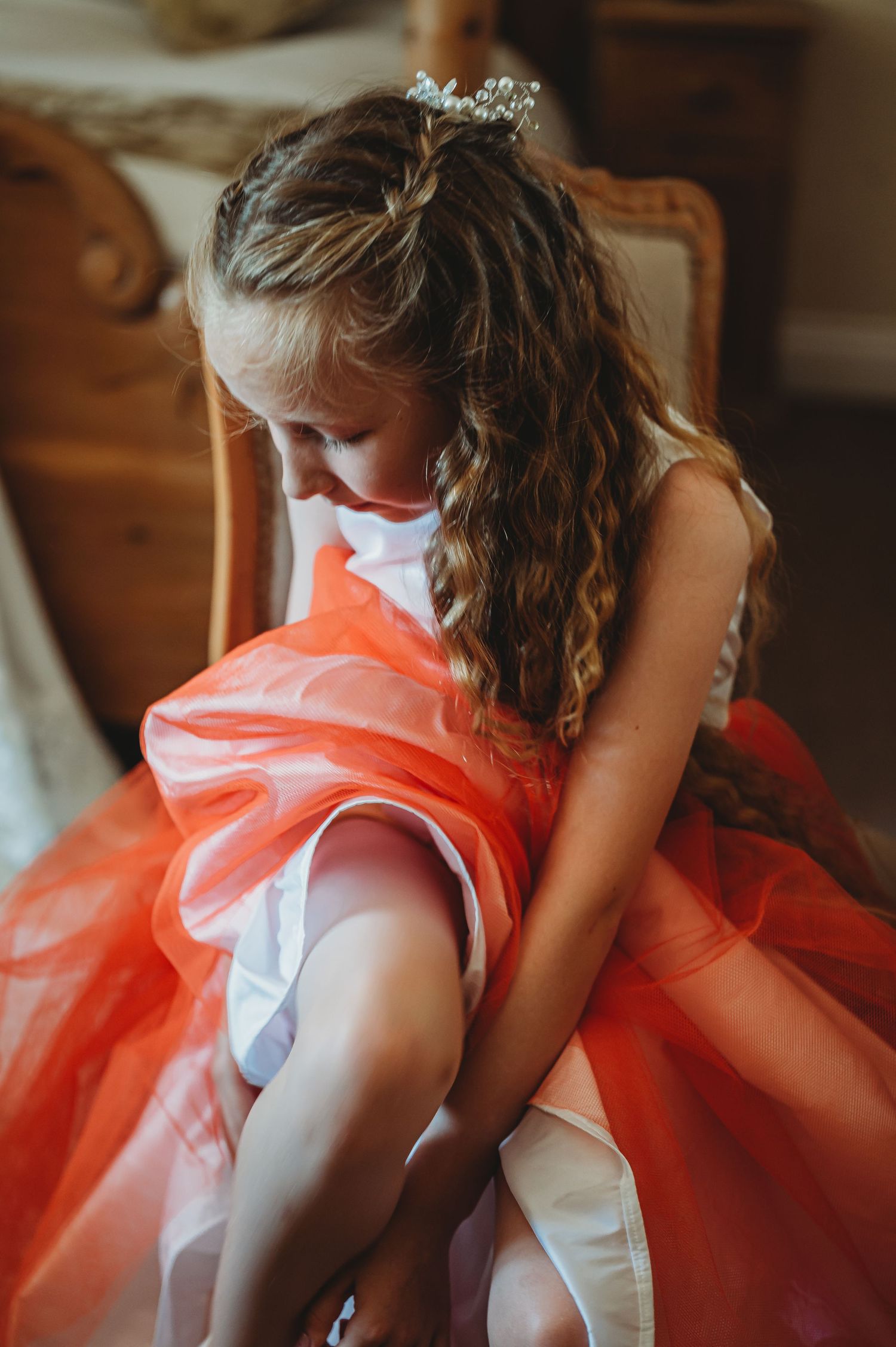A young dancer in a vibrant coral tulle dress adjusts her leg warmers while seated in an ornate wooden chair.