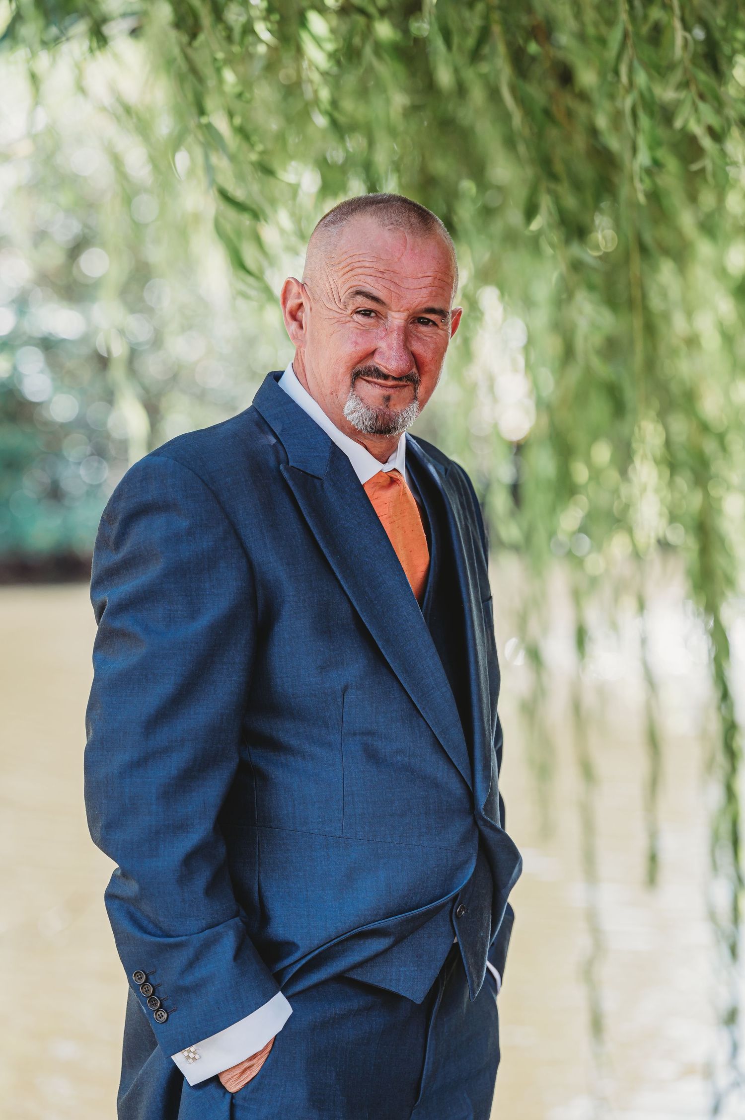 A person in a navy blue suit with orange tie stands confidently under drooping willow tree branches outdoors.