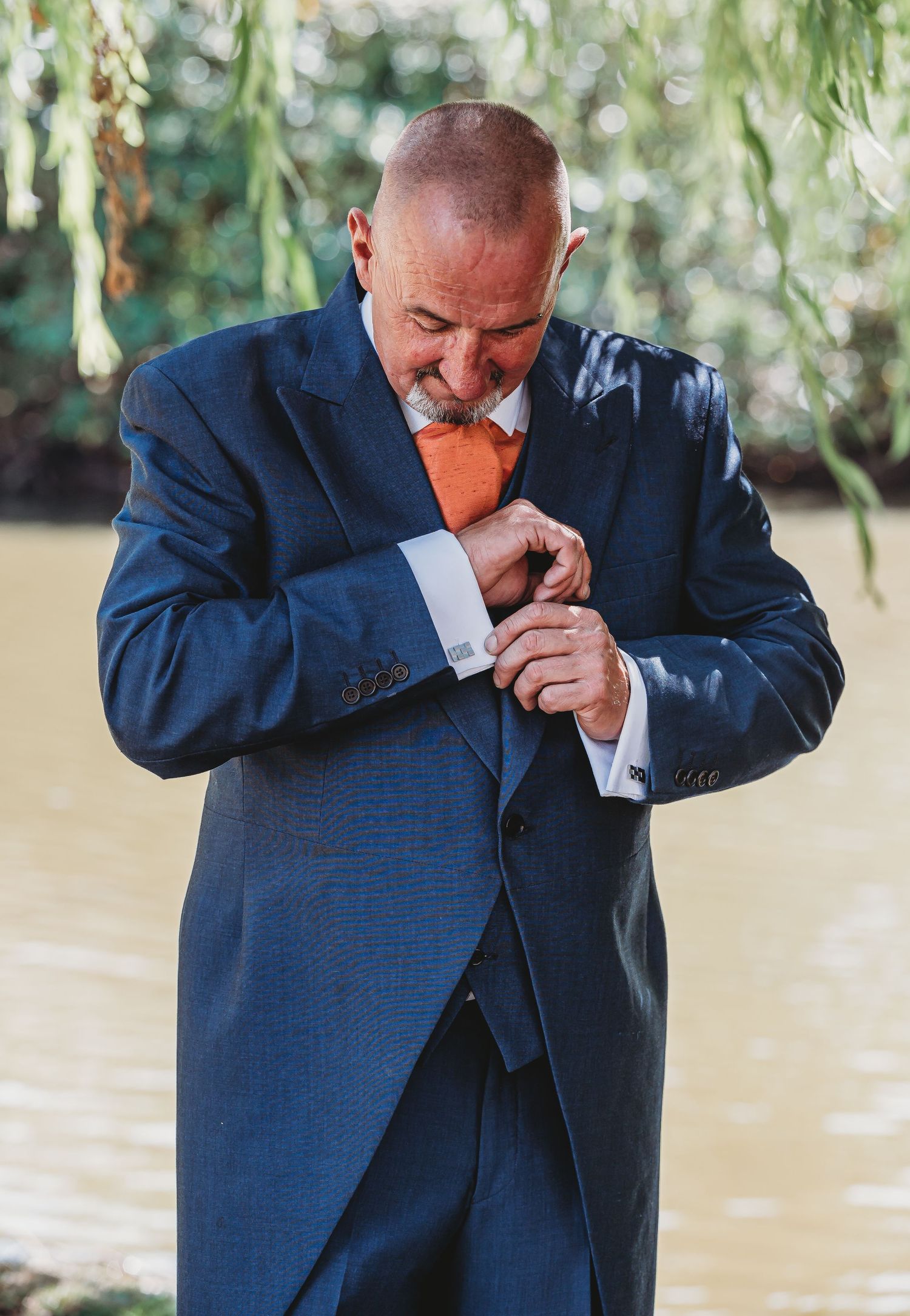 A person in a navy blue suit adjusts their sleeve cuff while smiling under willow tree branches outdoors.