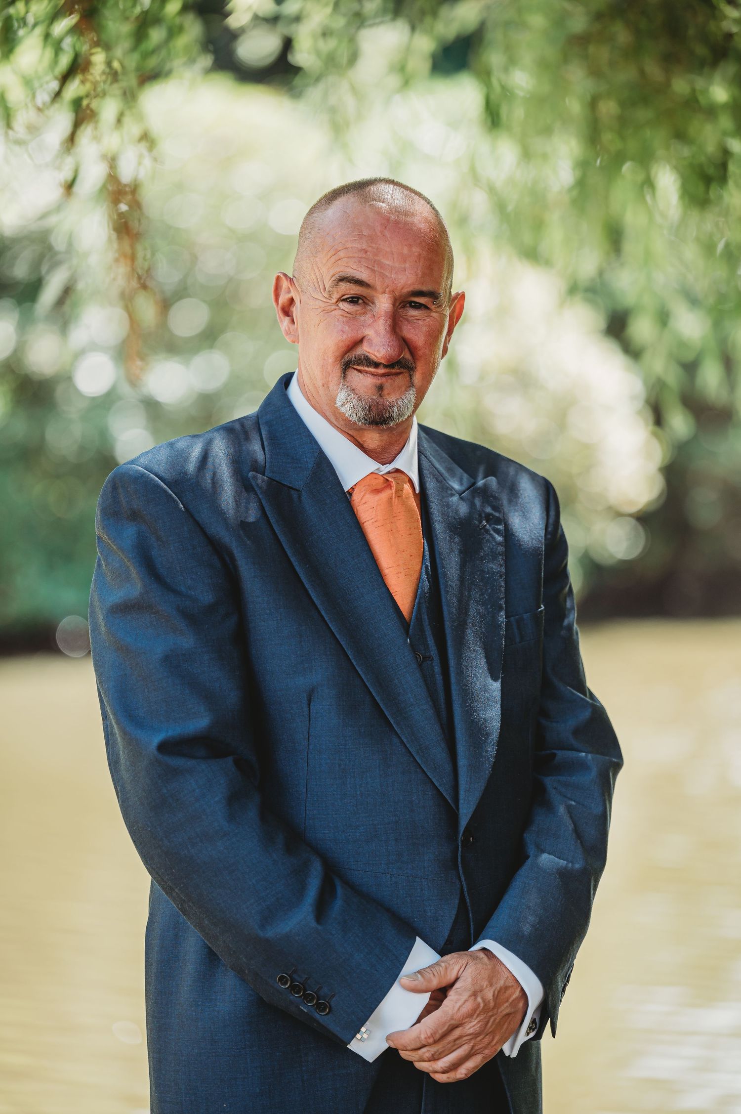 Professional portrait of someone in a navy suit with orange tie standing outdoors with soft natural lighting.