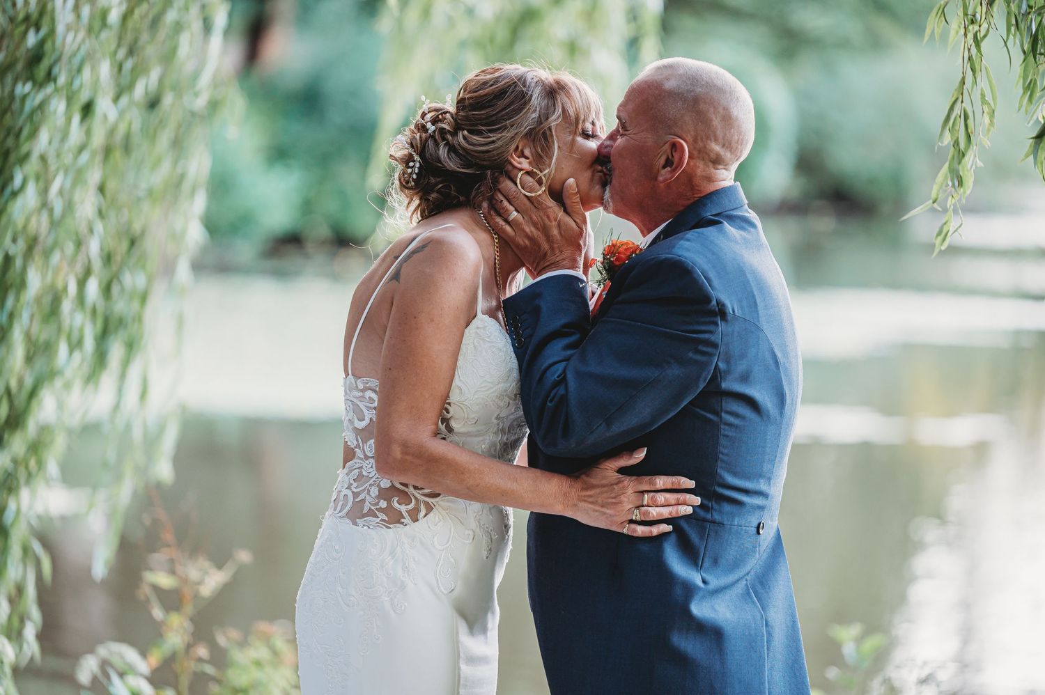 A romantic summer wedding kiss under weeping willow branches beside a serene pond.