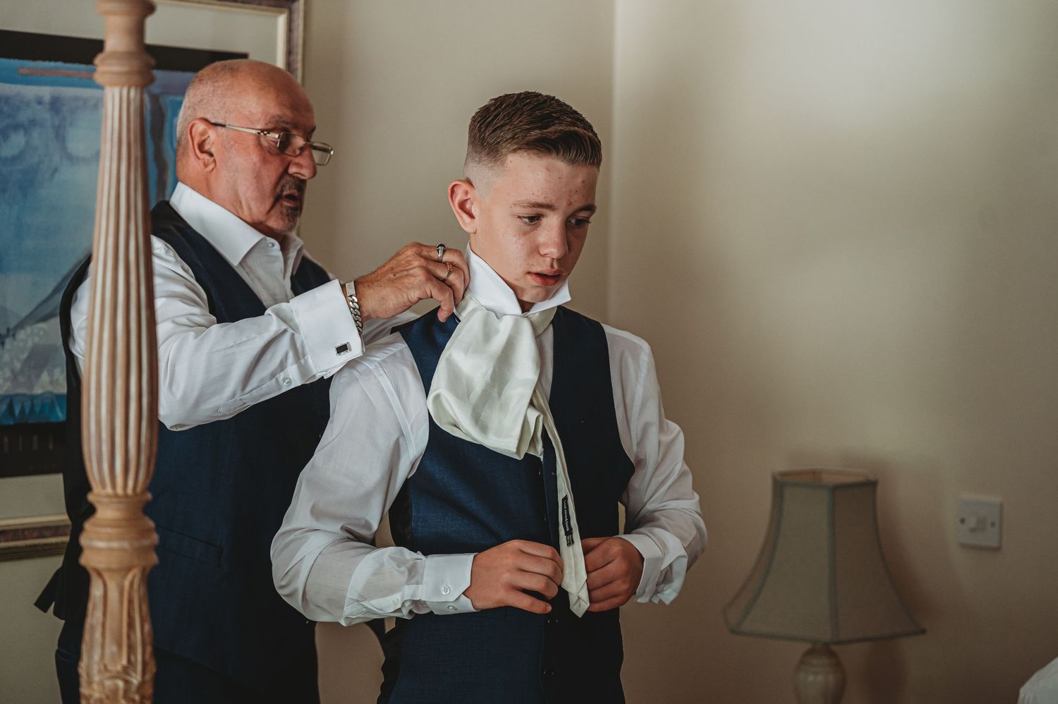 Two people in formal attire prepare for an event as an older gentleman adjusts a younger person's tie in a room.