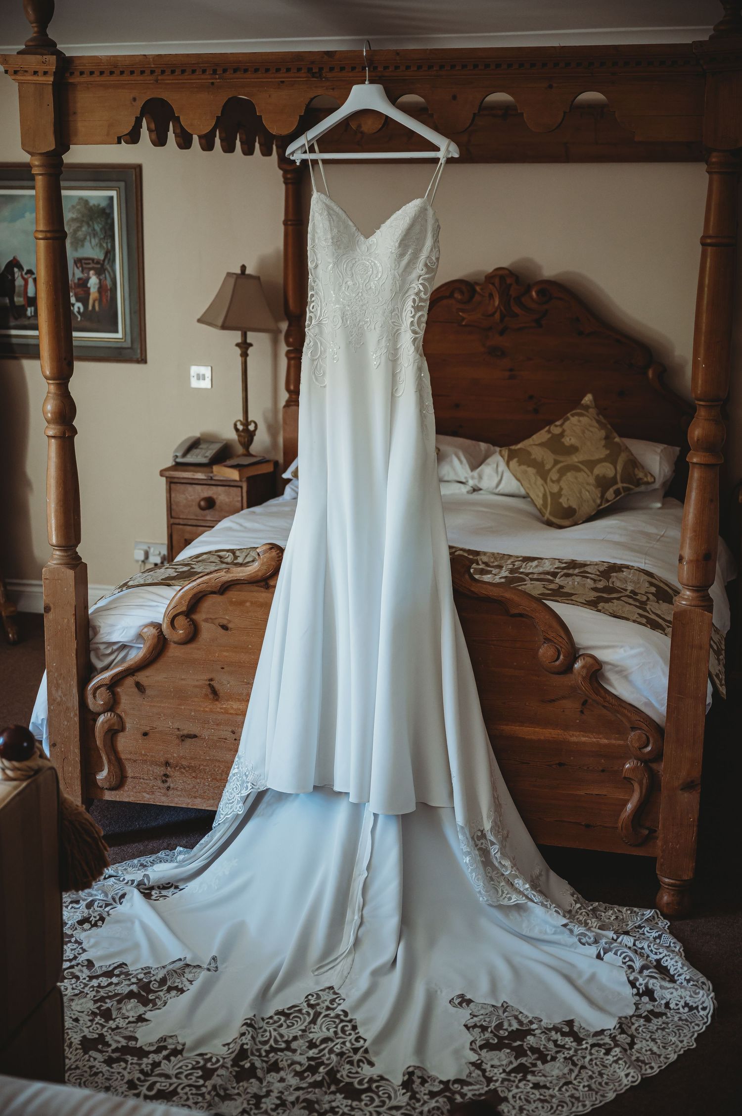 A elegant white wedding dress with lace trim and long train hanging on display against an antique wooden four-poster bed.
