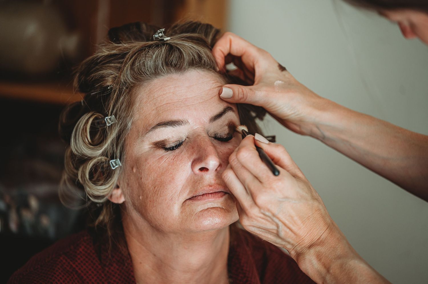 Makeup artist applies eyeliner while client sits with hair in curlers during beauty preparation.